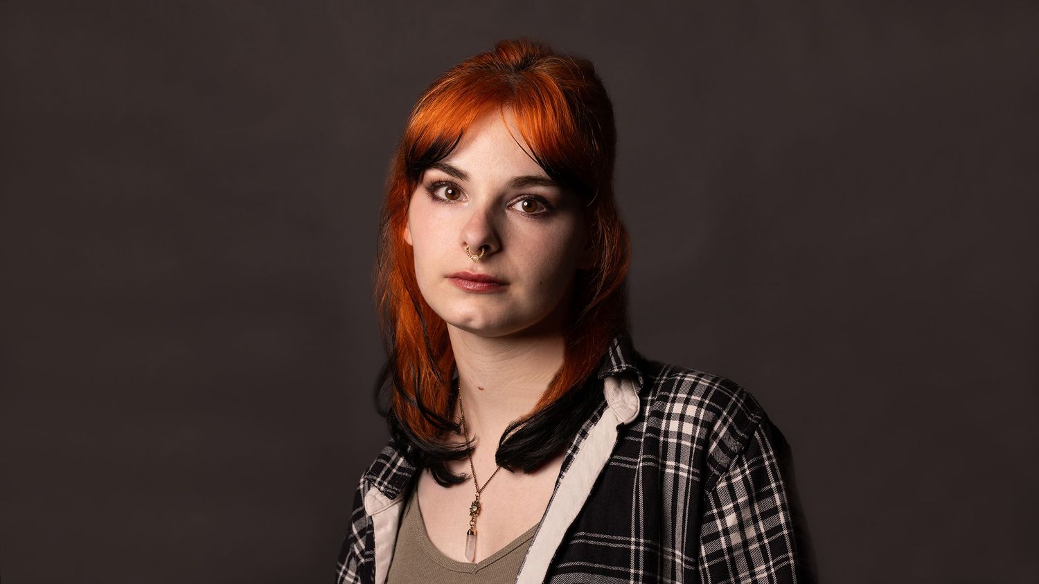 Young woman with bright orange shoulder-length hair wearing a black and white plaid jacket over a gray top against a dark studio backdrop.