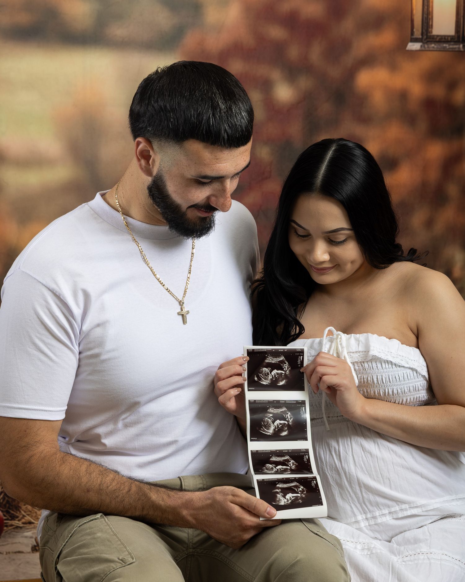 Couple holding ultrasound photos during maternity photoshoot with autumn backdrop.