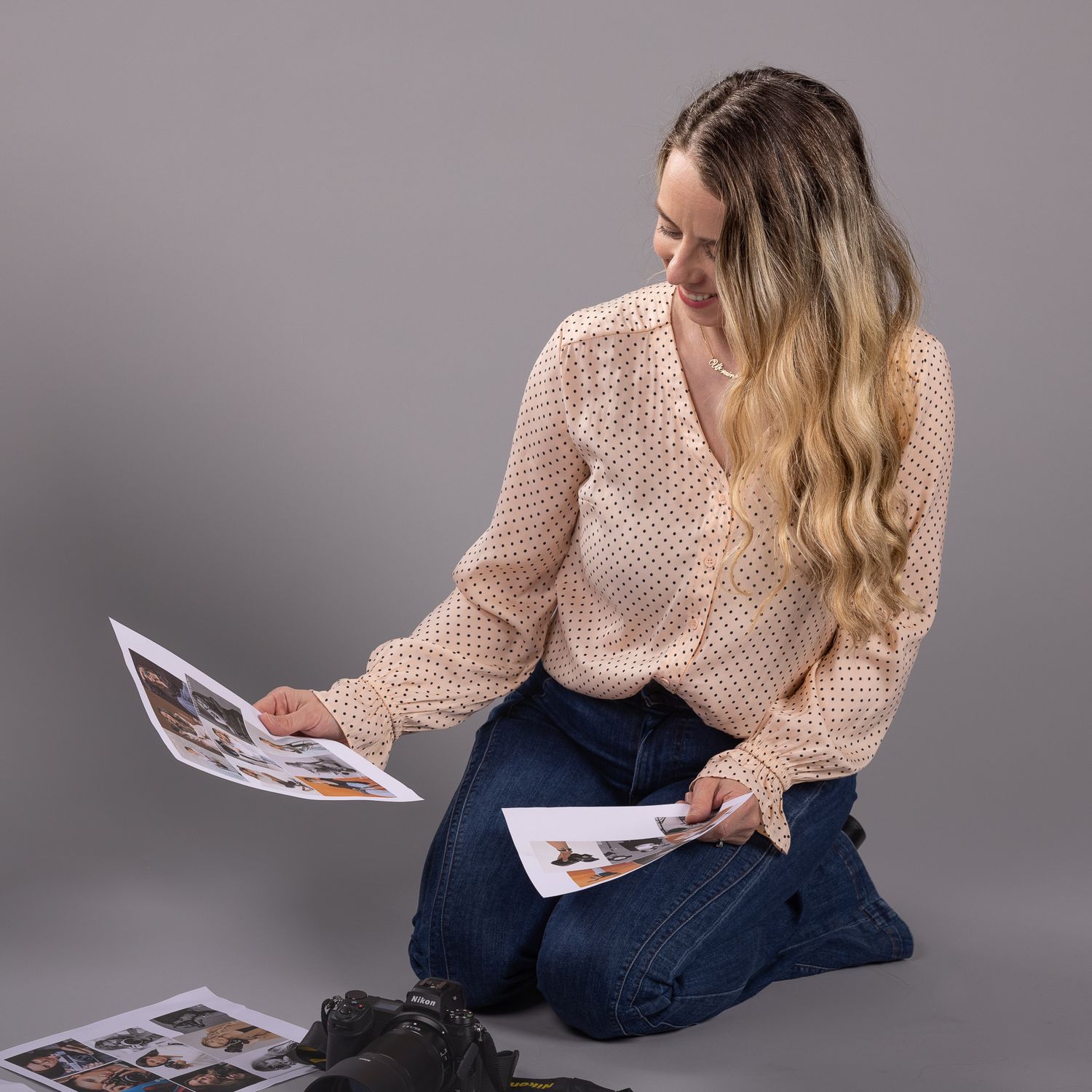 Woman in beige blouse and jeans sitting on floor reviewing printed photos spread around her.