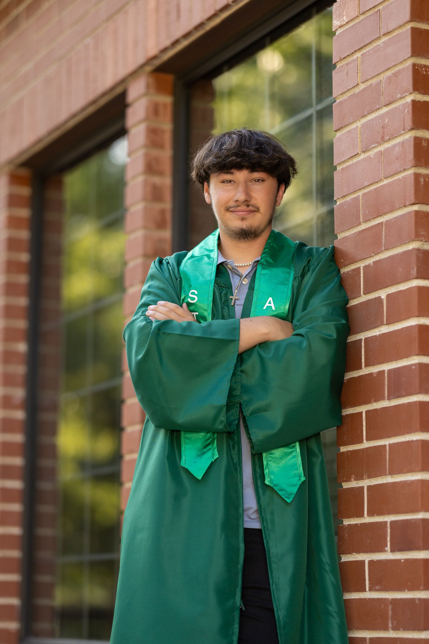 Young man in green graduation gown and stole poses confidently against brick wall outdoors.