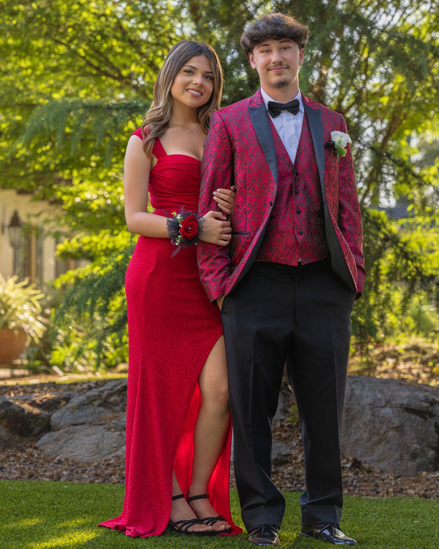 A couple dressed in matching red prom attire poses outdoors, she in a gown and he in a floral suit.