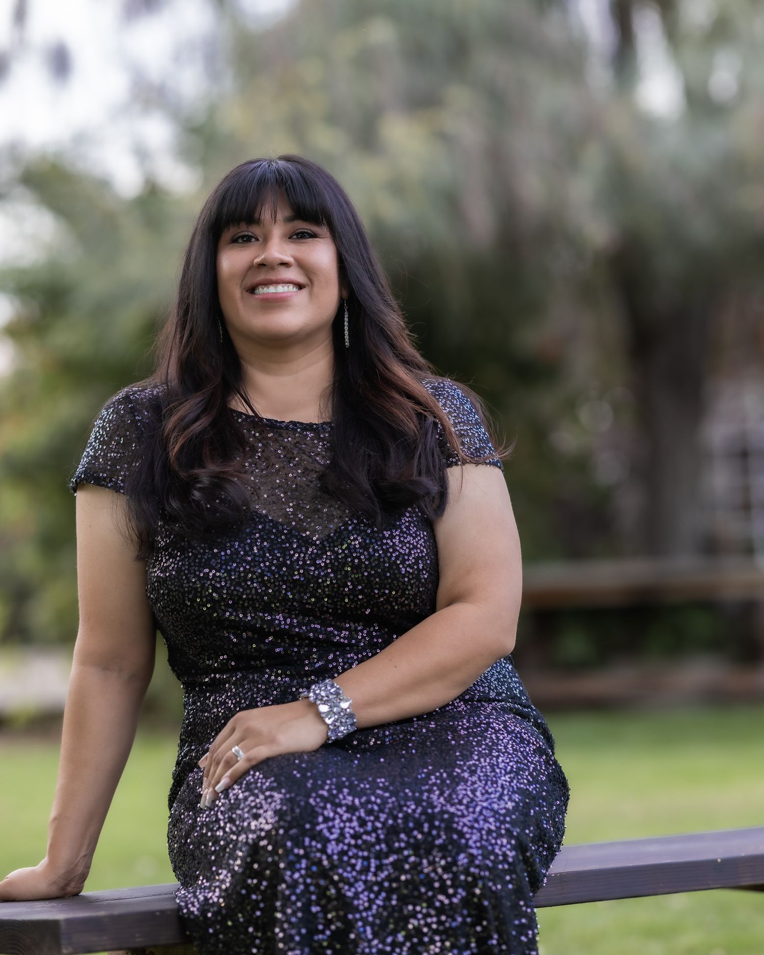 A smiling woman in a black sparkly dress poses outdoors on a wooden bench with lush green trees in the background.