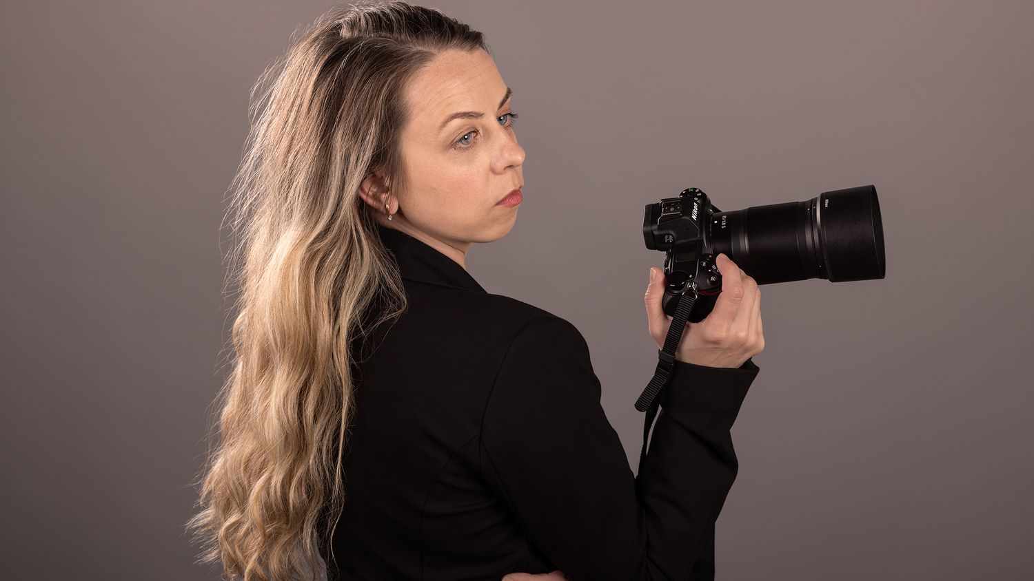 A blonde woman in black holding a DSLR camera with a telephoto lens, posing against a gray background.