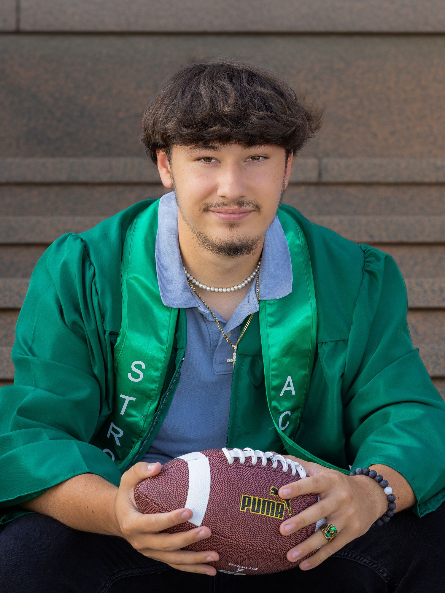 Young man in green graduation gown holding a Puma football, smiling for senior portrait photos outdoors.