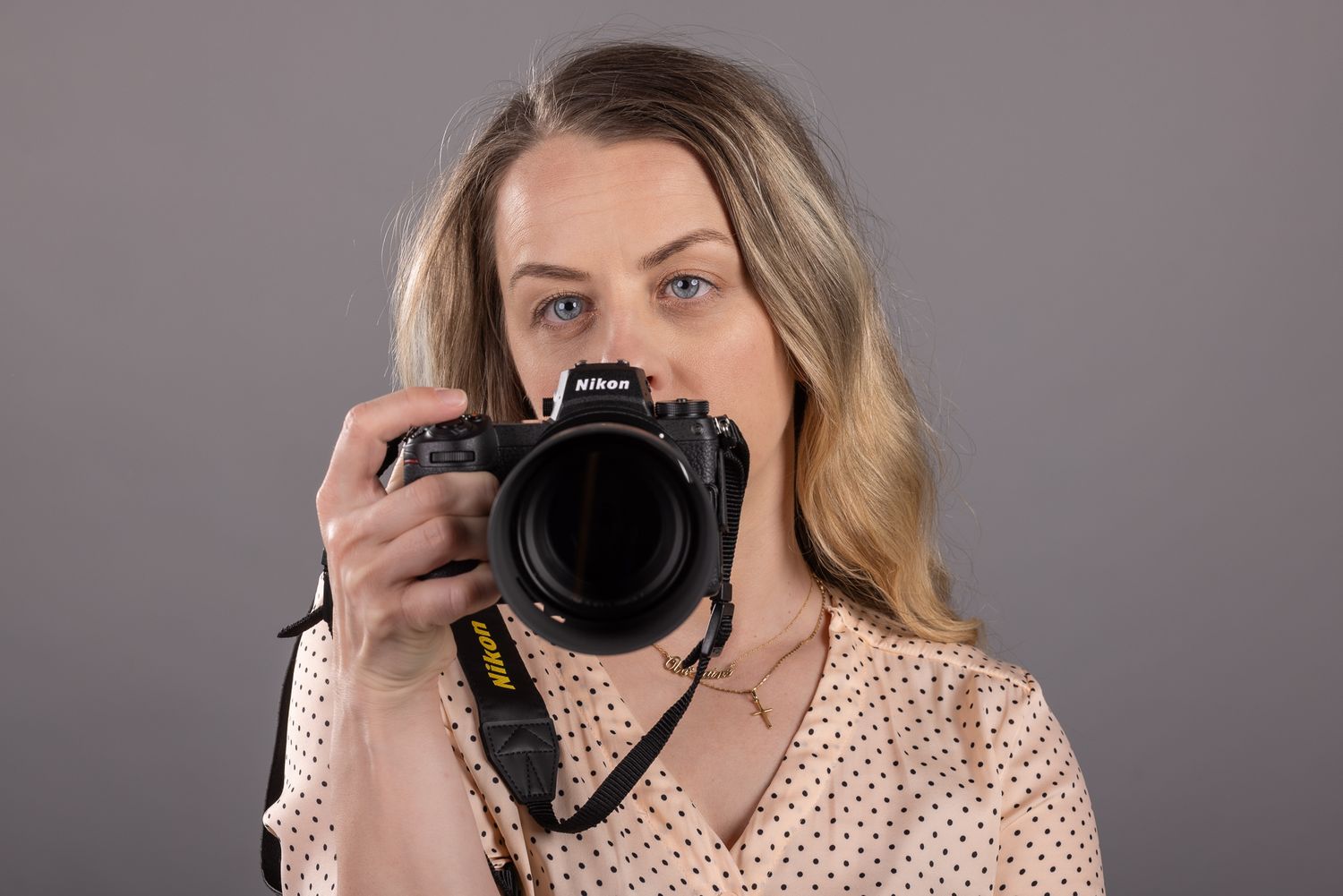 Blonde woman holding a Nikon DSLR camera up to her eye, photographing against a gray studio background.