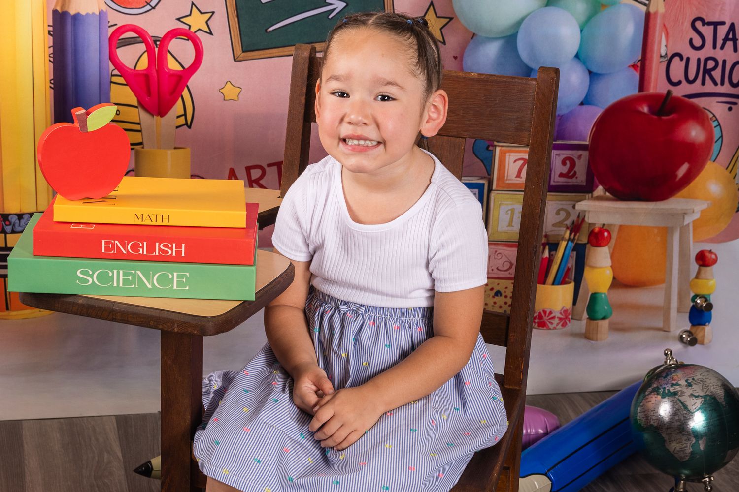 Young girl in white top and blue skirt smiling at back-to-school photo shoot with books and apple props.