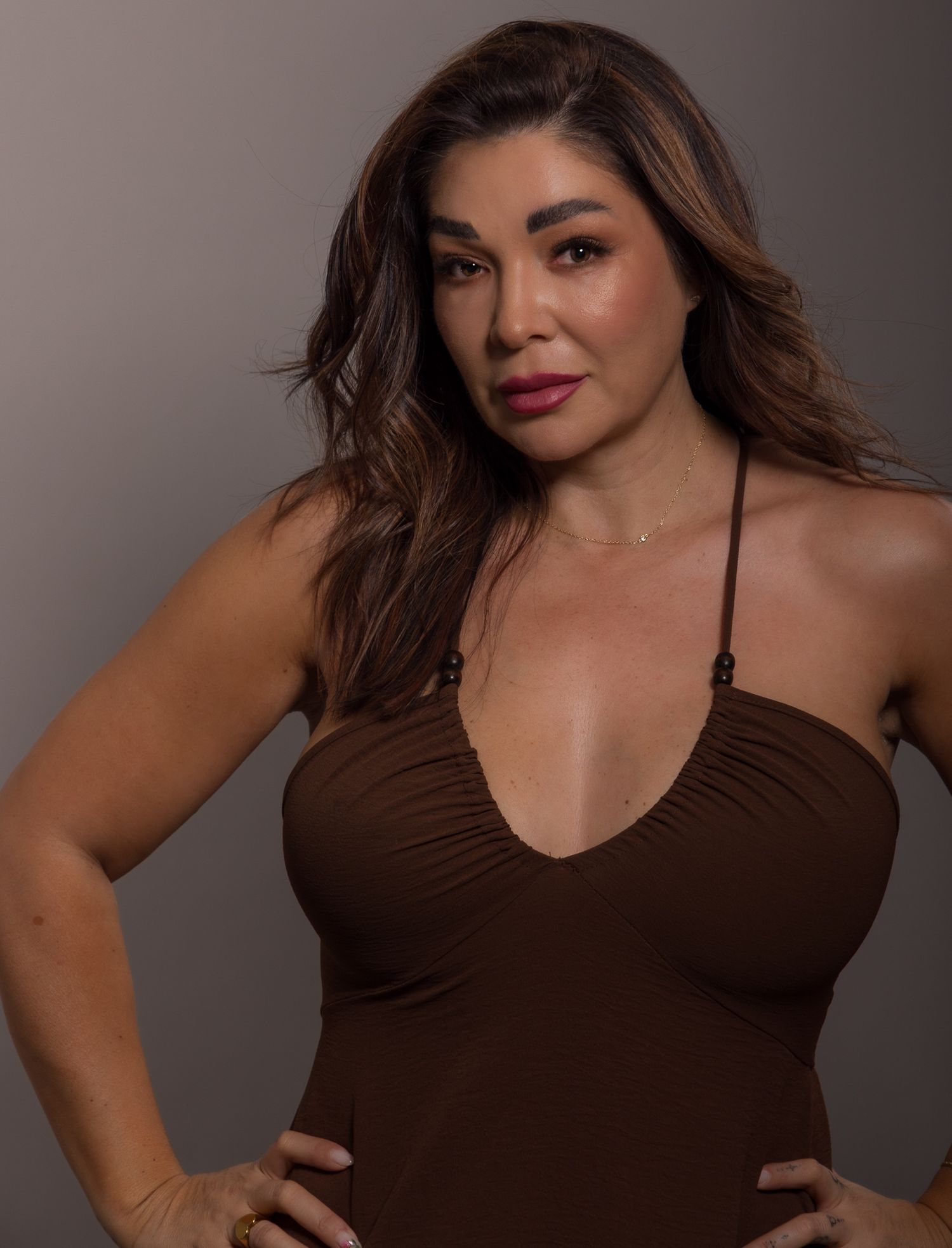 Woman in brown halter top posing confidently with hands on hips against a neutral gray background.