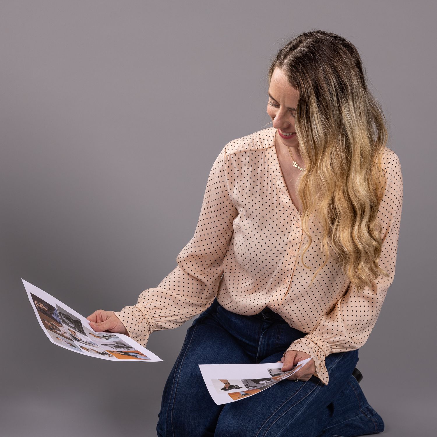 Young woman with long blonde hair sitting and reviewing printed photos on gray studio background.