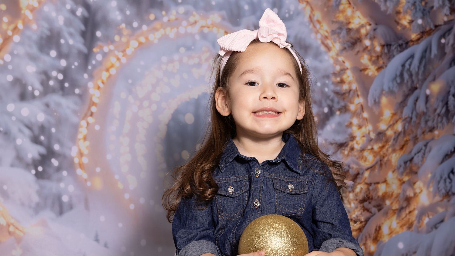 Smiling young girl with a pink bow and denim jacket holds a gold ornament against a sparkly snowy winter holiday backdrop.