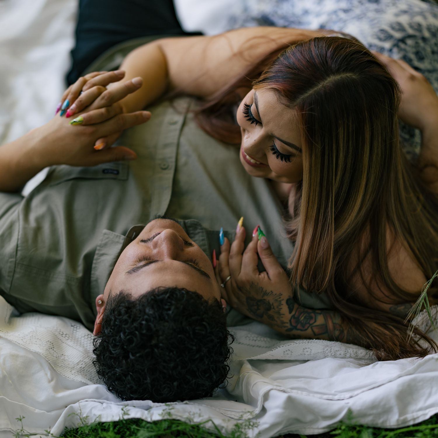 A couple lying together on a white bed, smiling and holding hands with colorful nails and tattoos visible.