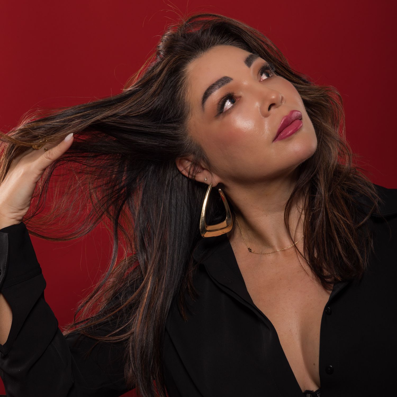 A woman in a black top with gold earrings poses against a red background, tossing her dark hair dramatically.