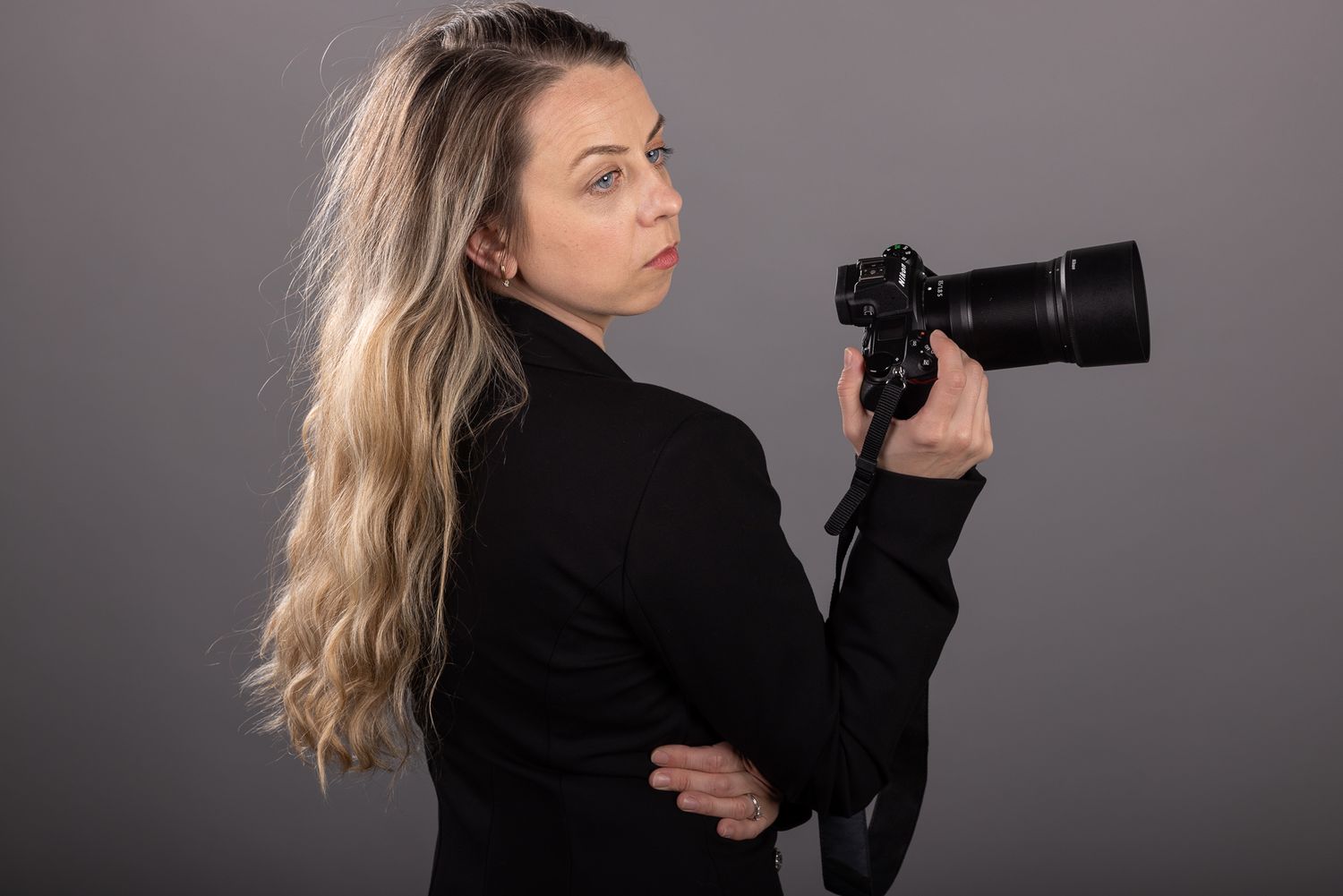 Blonde woman in black blazer holding a professional DSLR camera with telephoto lens against gray background.