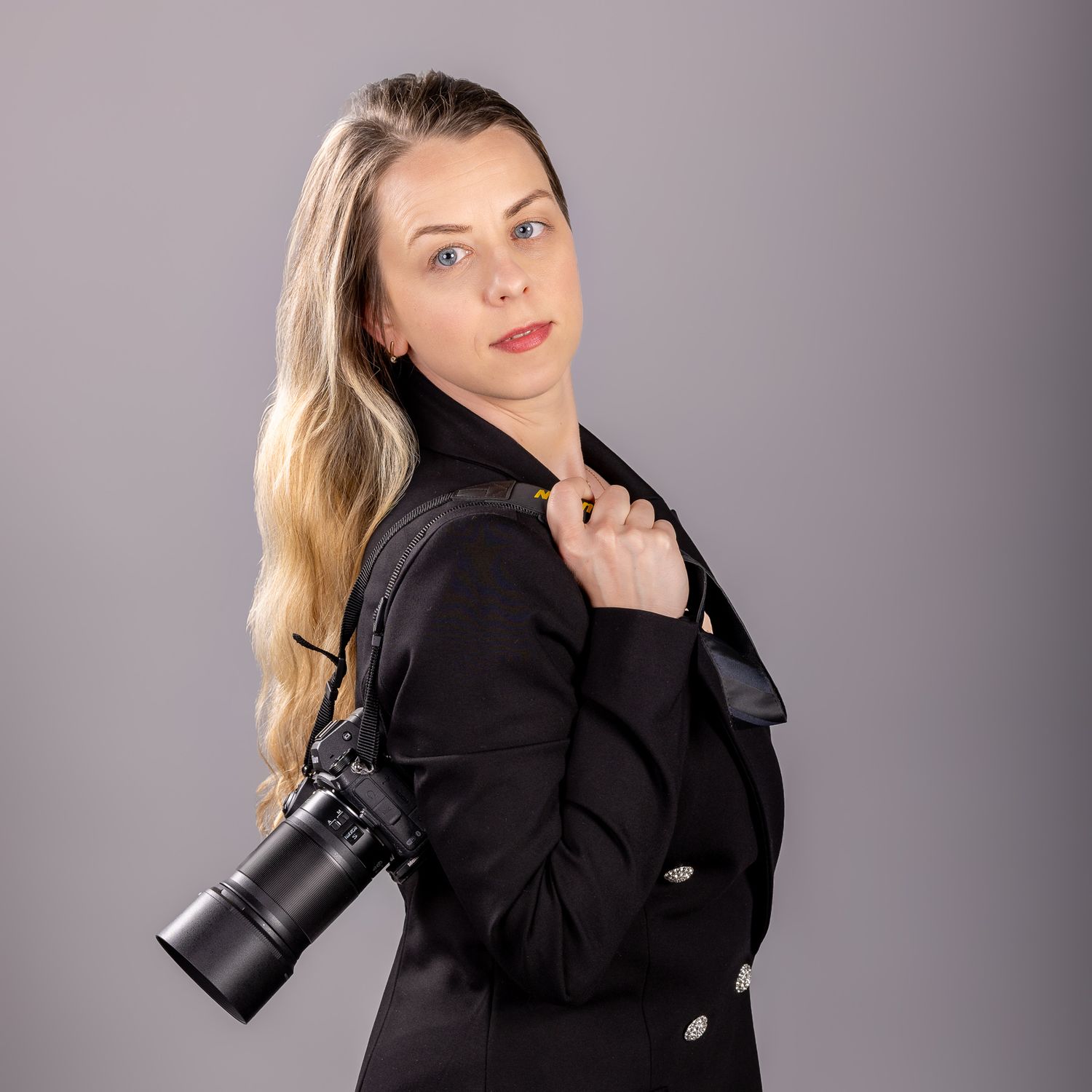 Blonde woman in black jacket holding a professional camera over her shoulder against a gray background.