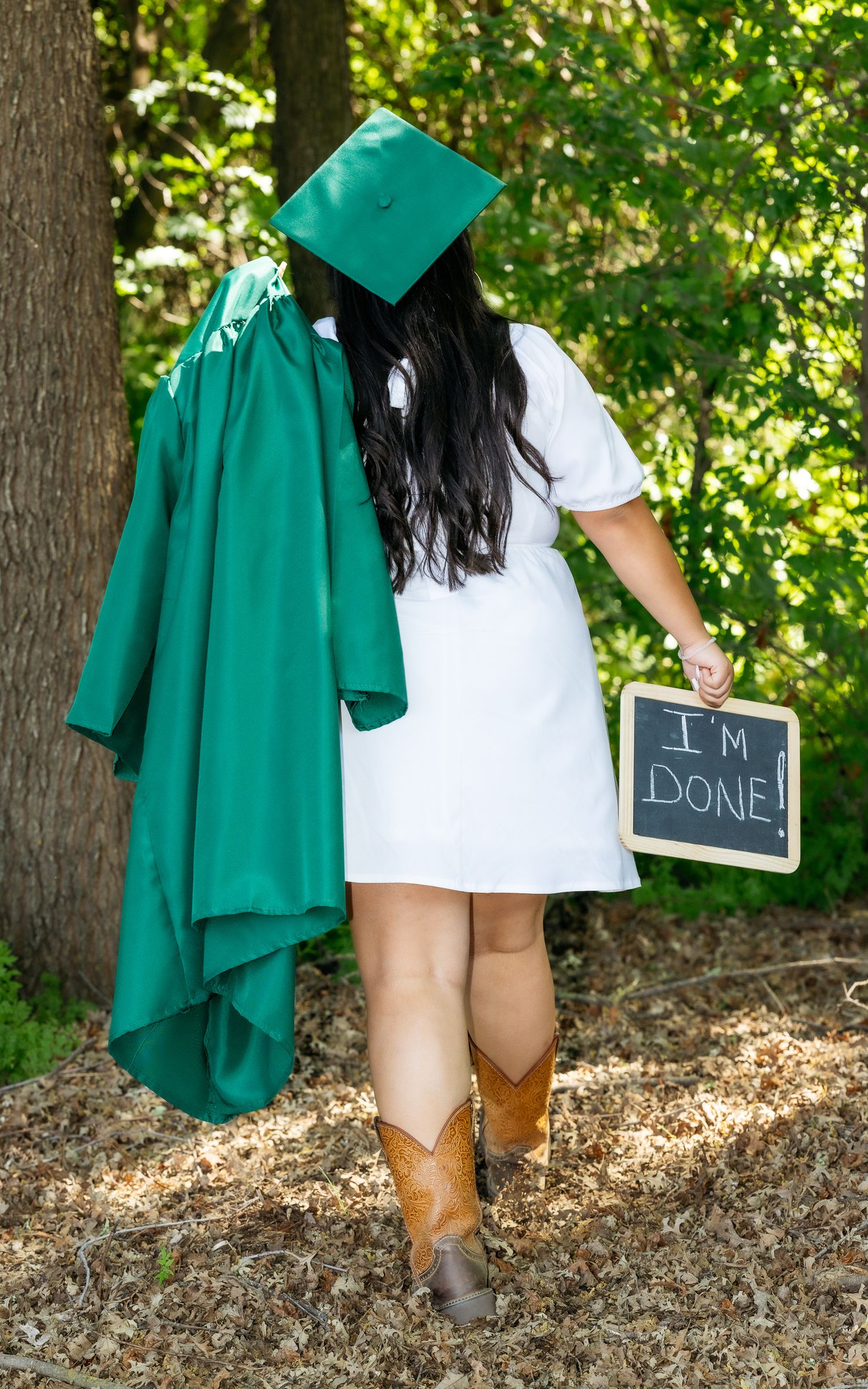Graduate holding green cap and gown with 'I'm Done' chalkboard sign, wearing white dress and cowboy boots outdoors.