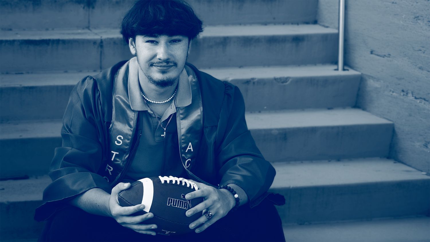 Young man sitting on concrete steps holding a football, wearing a jacket, in a blue-toned monochrome photo.