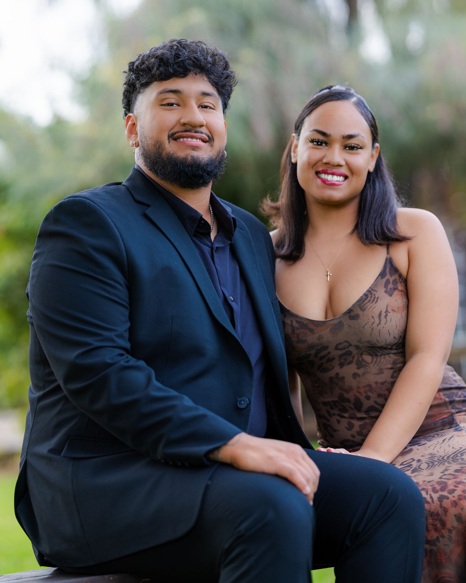A smiling couple in formal attire poses together outdoors, man in black suit and woman in brown lace dress.