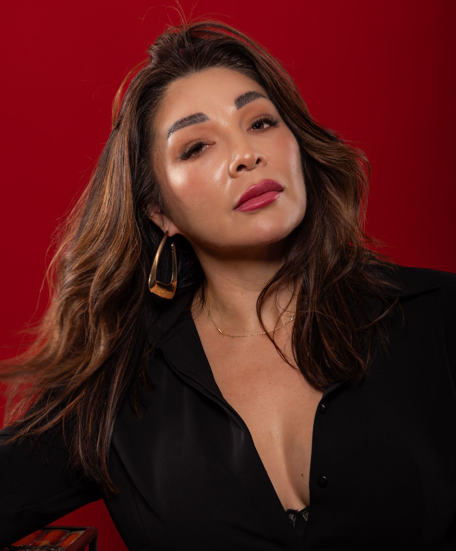 Woman with brown hair wearing gold hoop earrings and black top poses against red background.