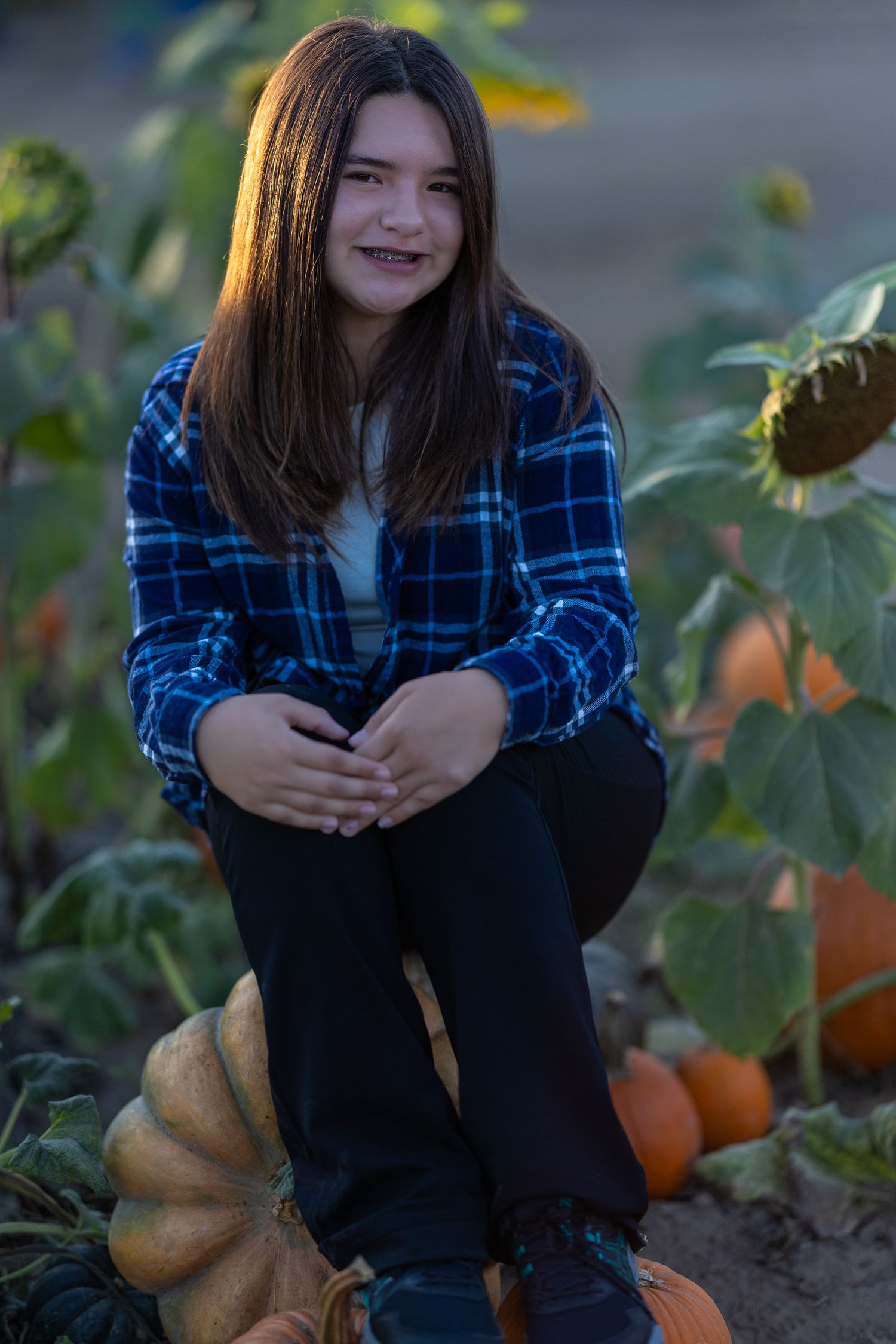 A smiling teenage girl in a blue plaid shirt sits on a pumpkin in a fall pumpkin patch with sunflowers.