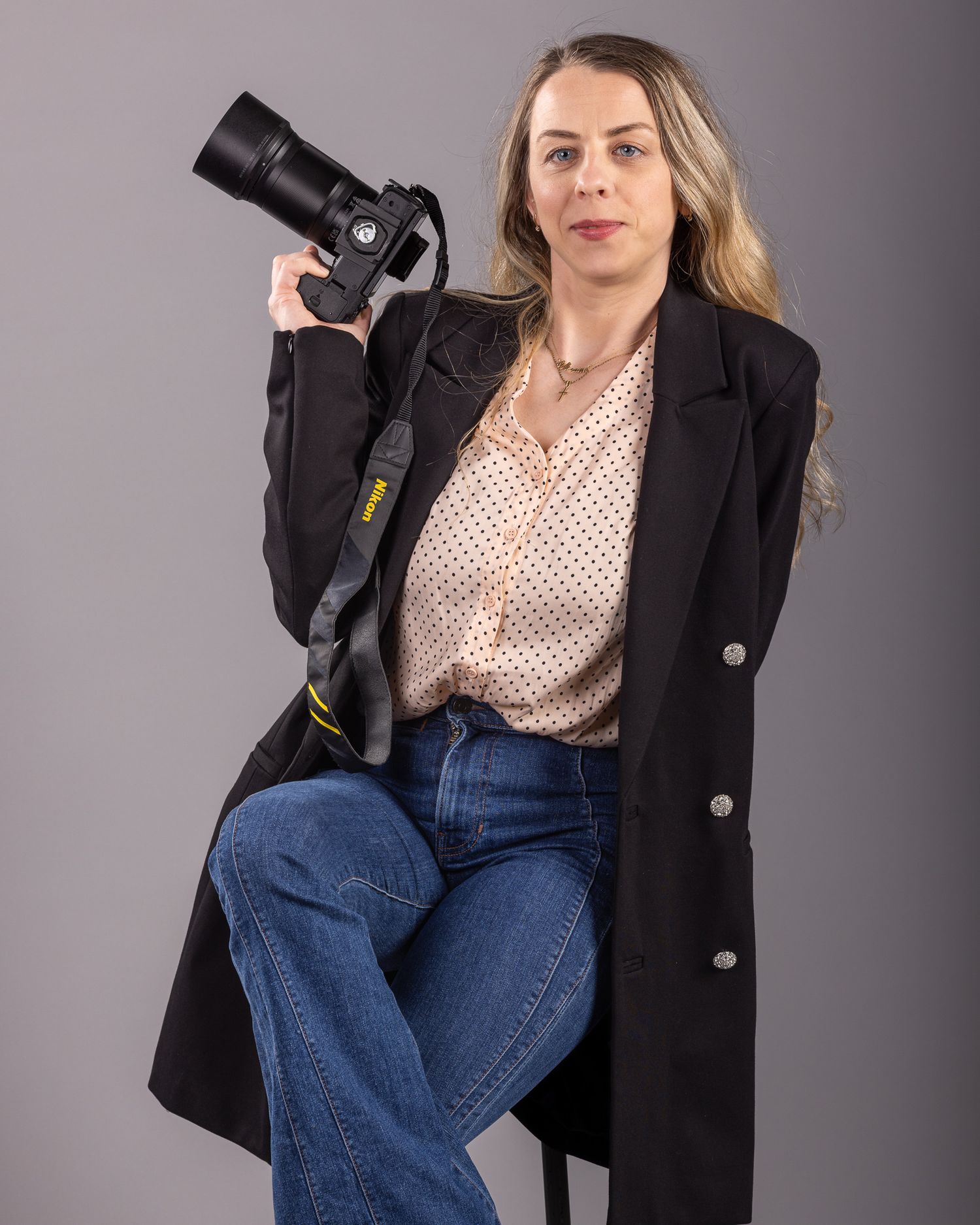 Woman photographer in black blazer and jeans poses holding a Nikon camera on gray studio background.