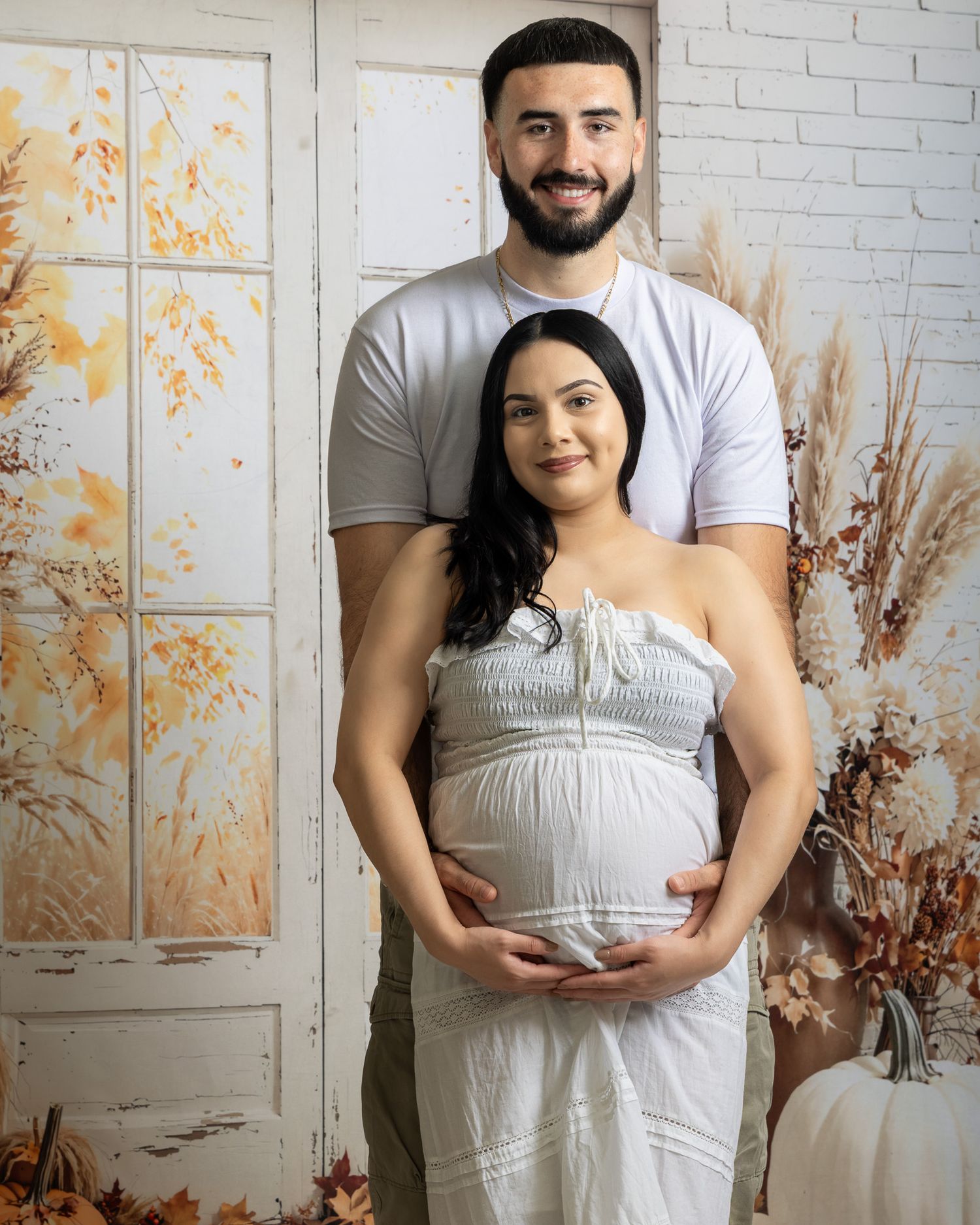 Couple posing for maternity photo shoot with fall-themed backdrop, woman in white dress cradling baby bump.
