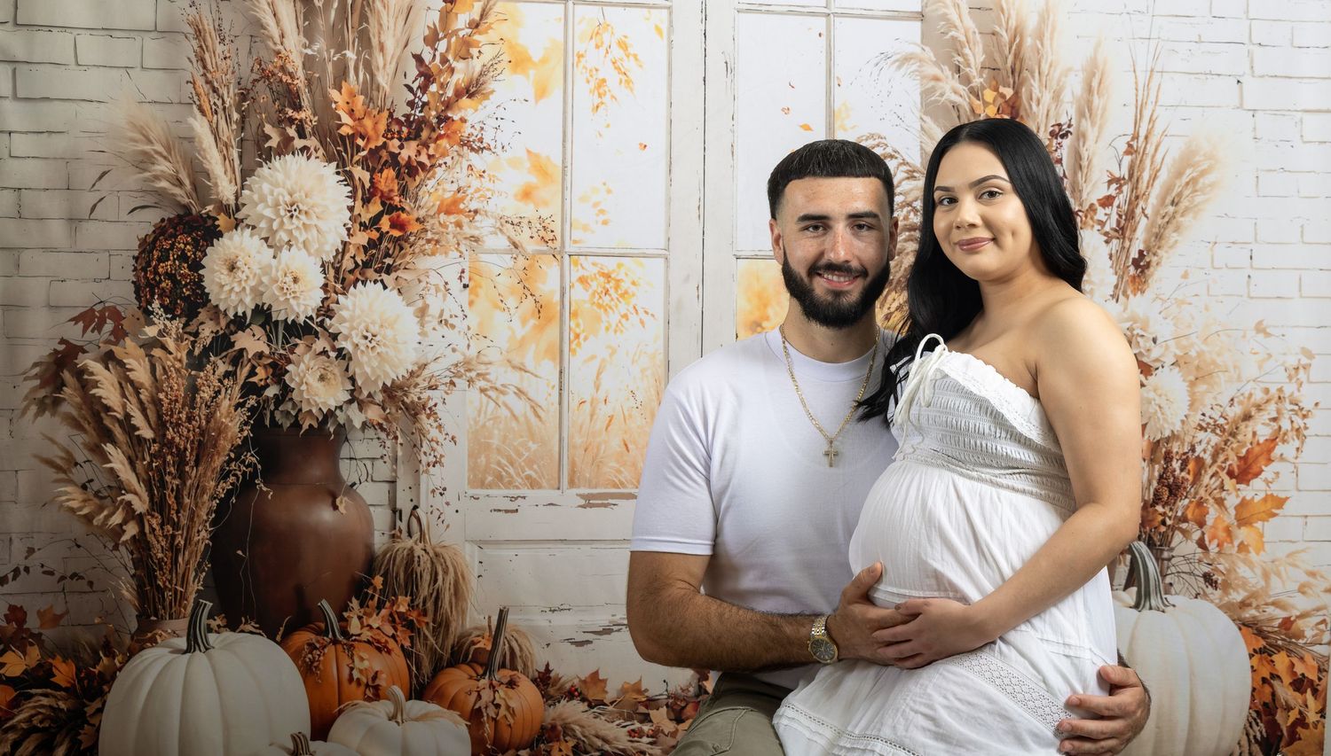 Couple poses for fall maternity photo with pumpkins and dried floral arrangements against a white backdrop.