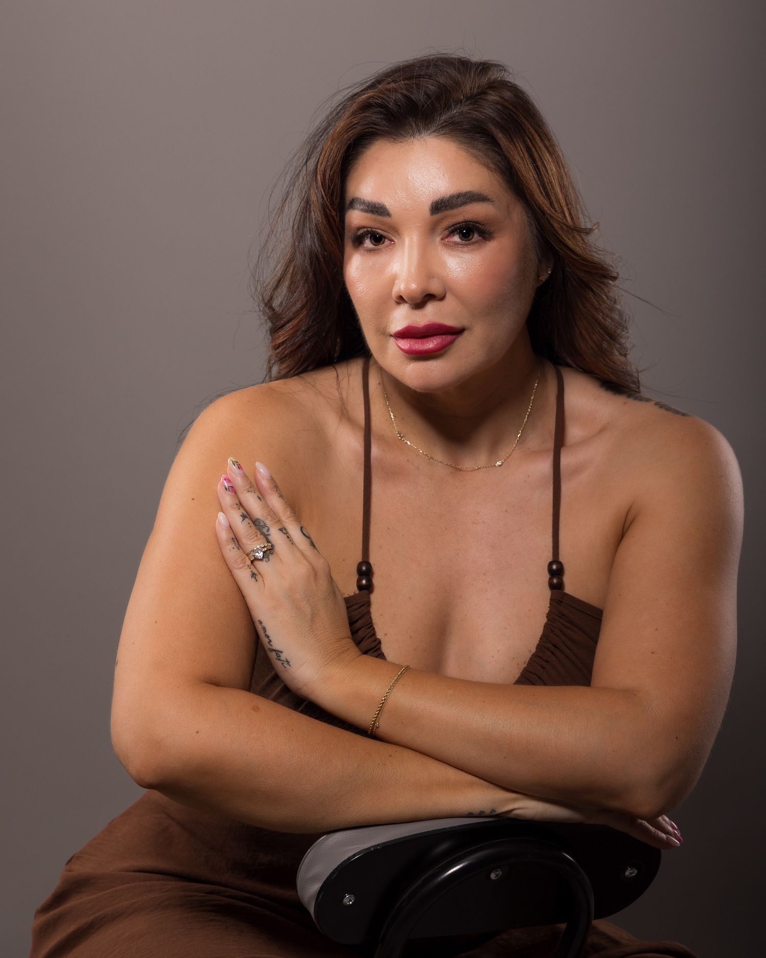 A woman in a brown spaghetti-strap top poses with arms crossed, wearing gold jewelry and rings against a gray background.