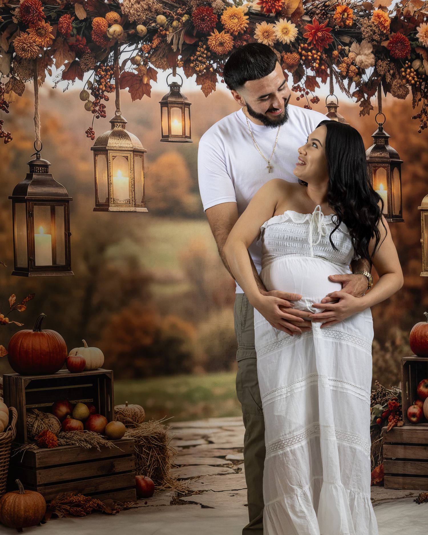 Couple poses for fall maternity photo with lanterns, pumpkins, and autumn floral arch in warm seasonal setting.