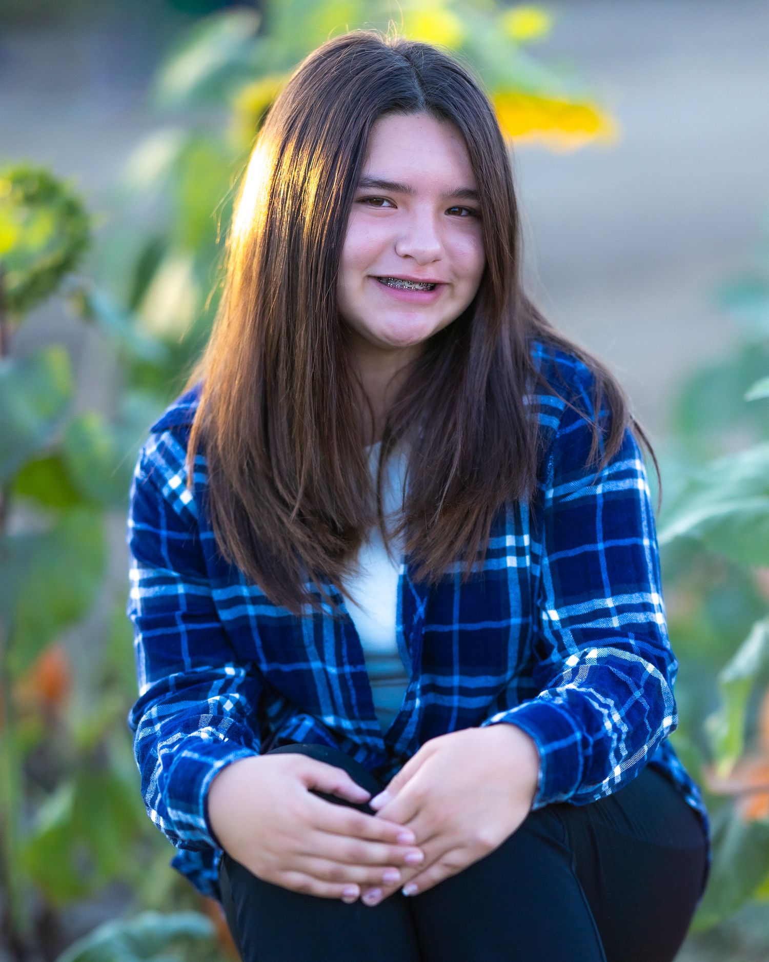 A smiling young girl with long brown hair wearing a blue plaid flannel shirt, sitting outdoors near sunflowers.