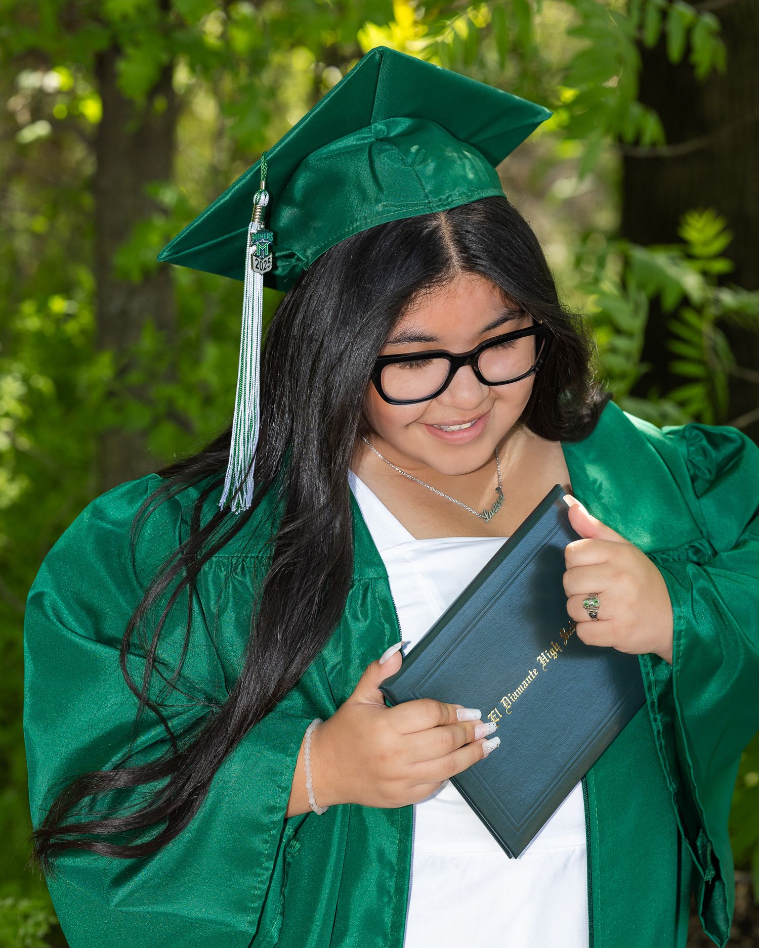 Smiling female graduate in green cap and gown holding diploma, wearing glasses outdoors.