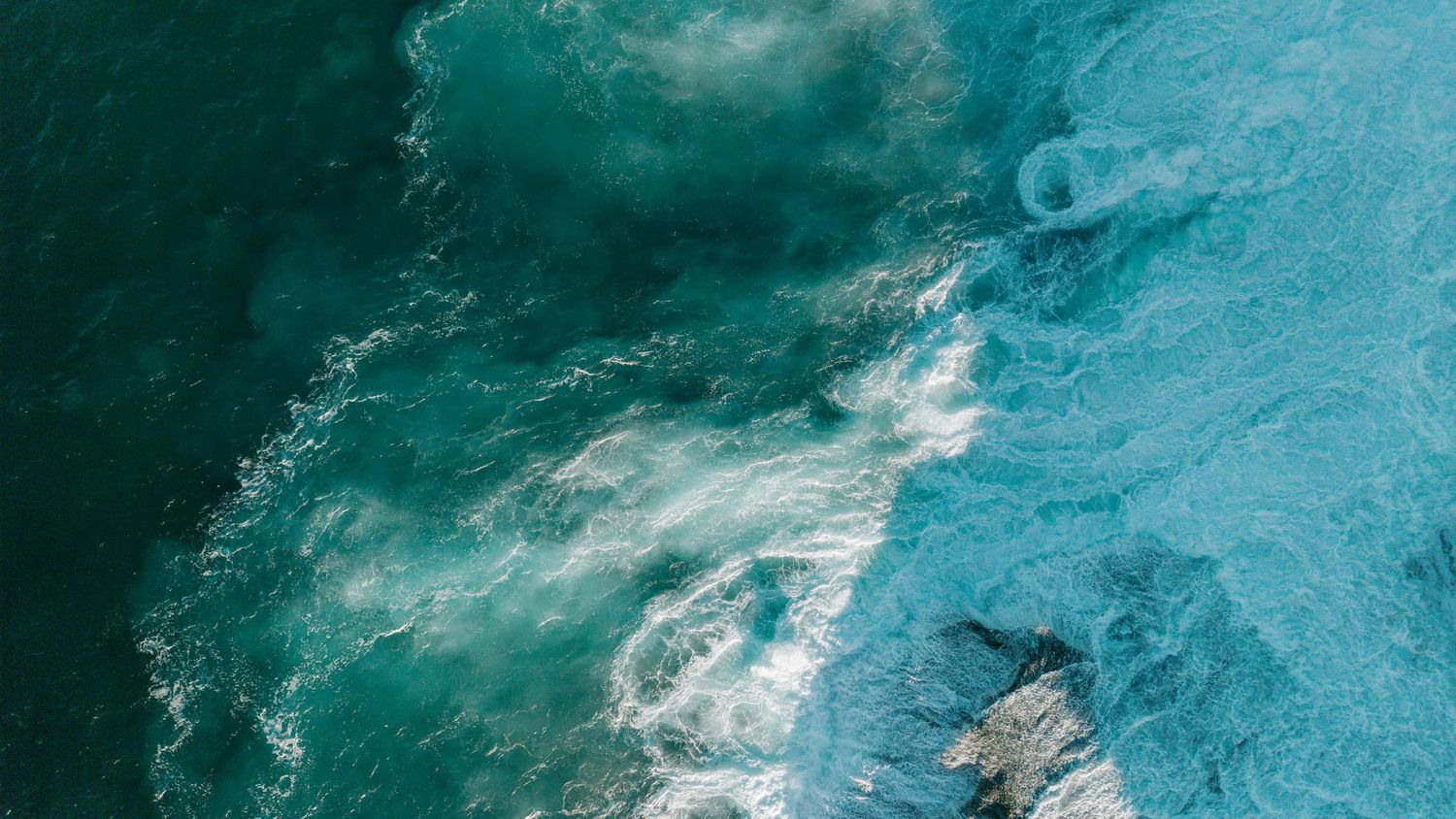 Aerial view of turquoise ocean waves crashing against a rocky coastline with swirling white foam patterns.