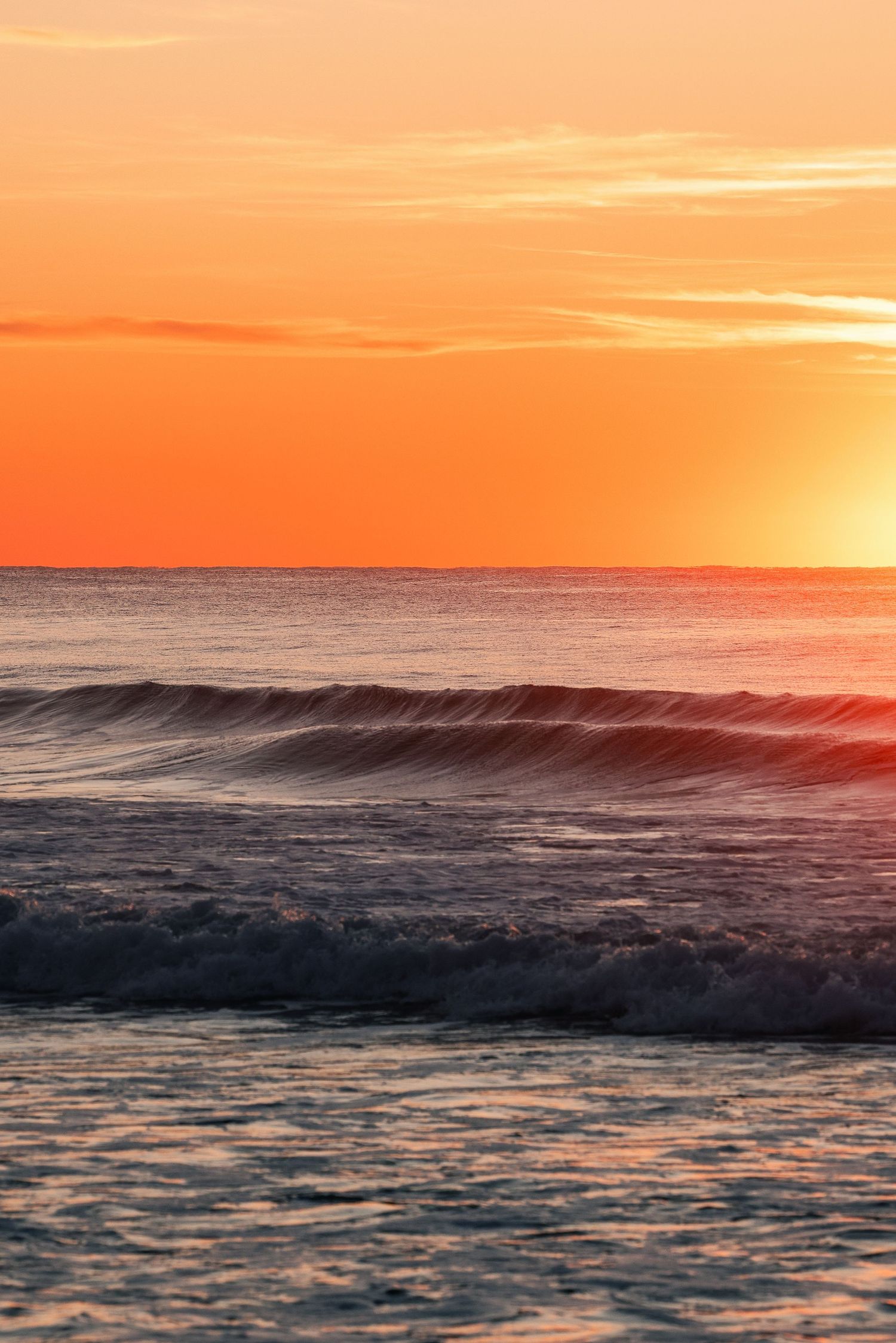 Ocean waves roll beneath a vibrant orange sunset sky with golden light reflecting on the water.