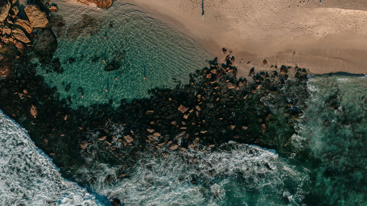 Aerial view of a rocky coastline with turquoise waves crashing onto a sandy beach surrounded by dark boulders.