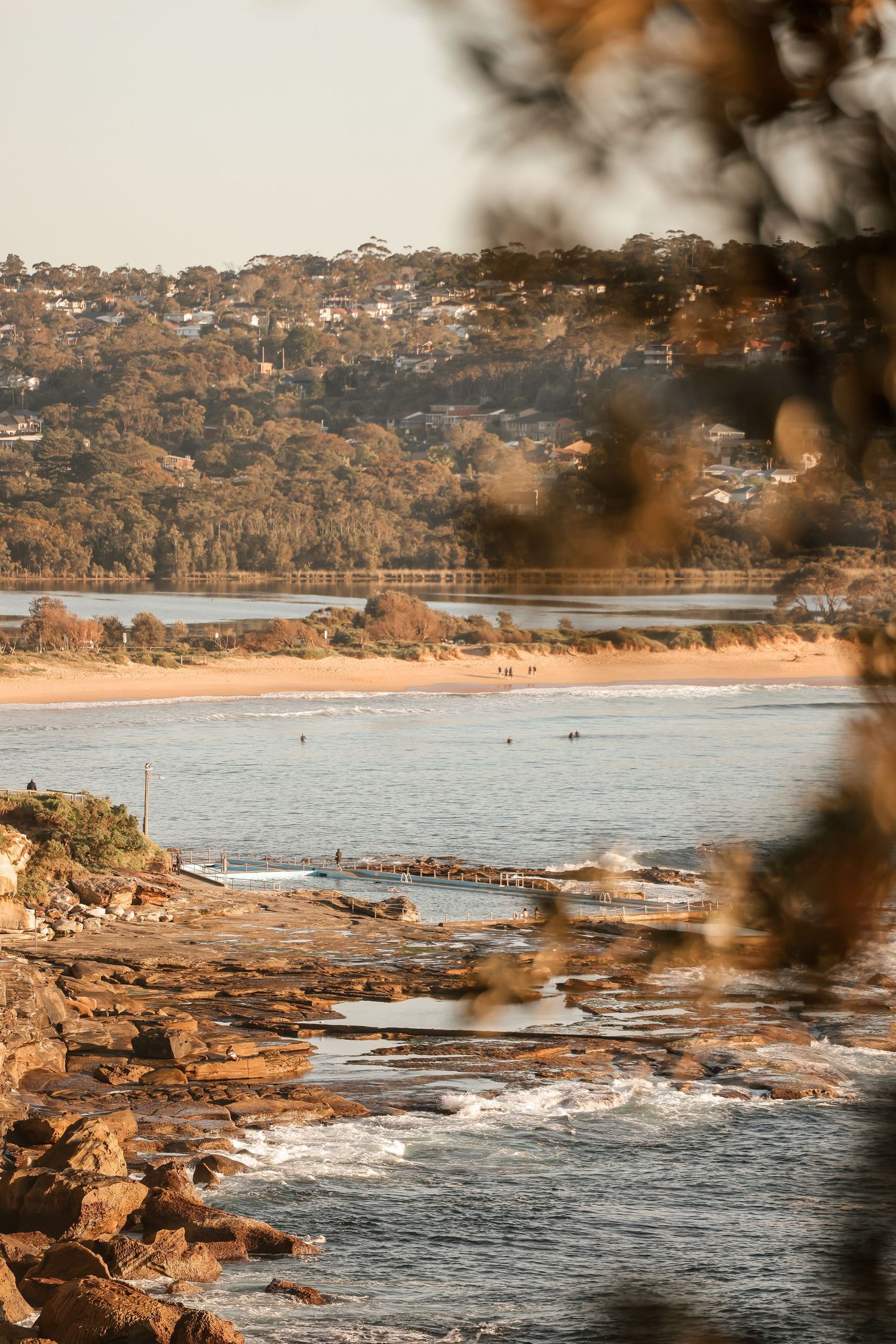 Rocky coastal shoreline with waves crashing against cliffs and beach houses visible in the distance.