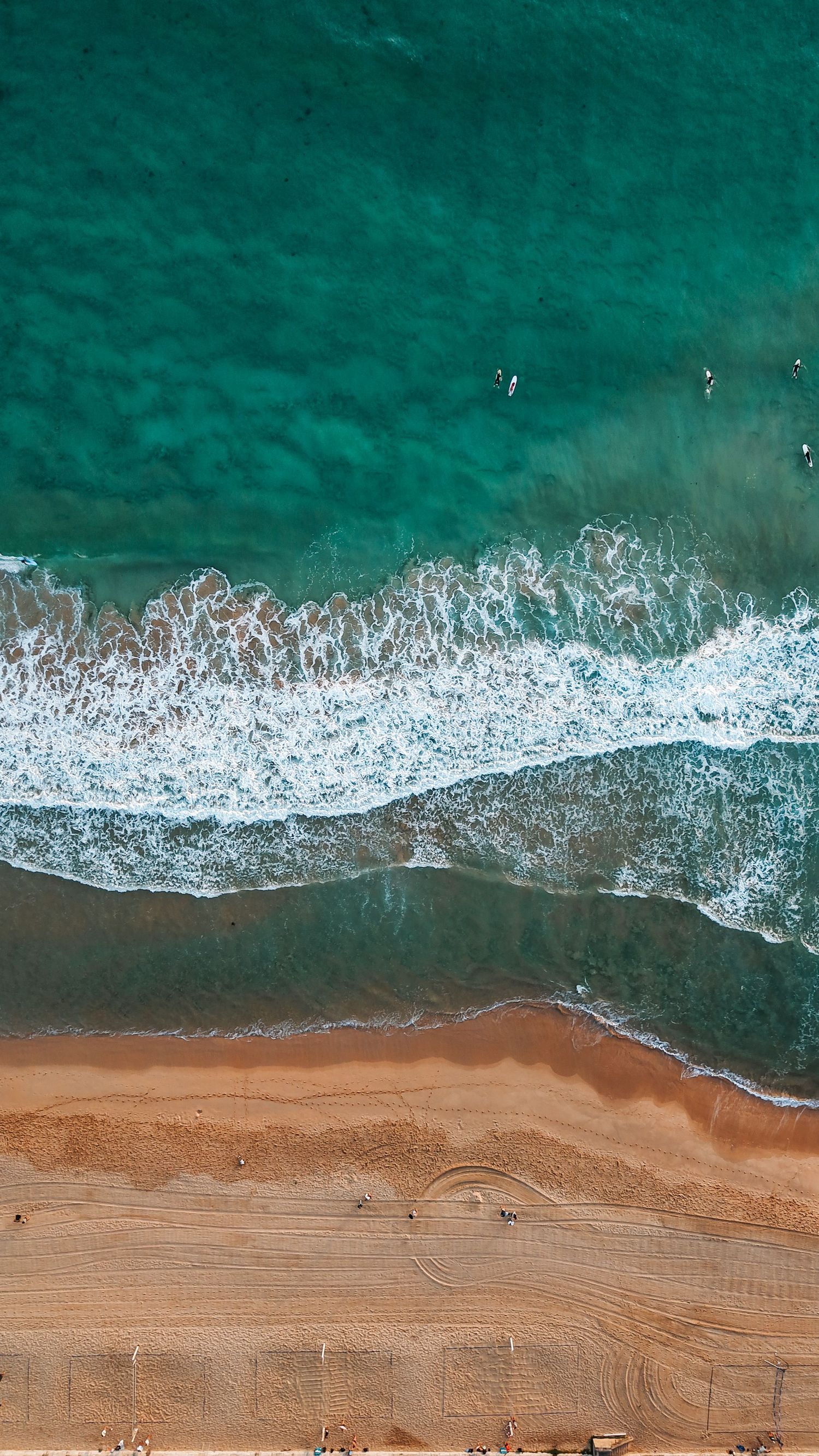 Drone shot of emerald waters crashing onto sandy beach creating white foam patterns.