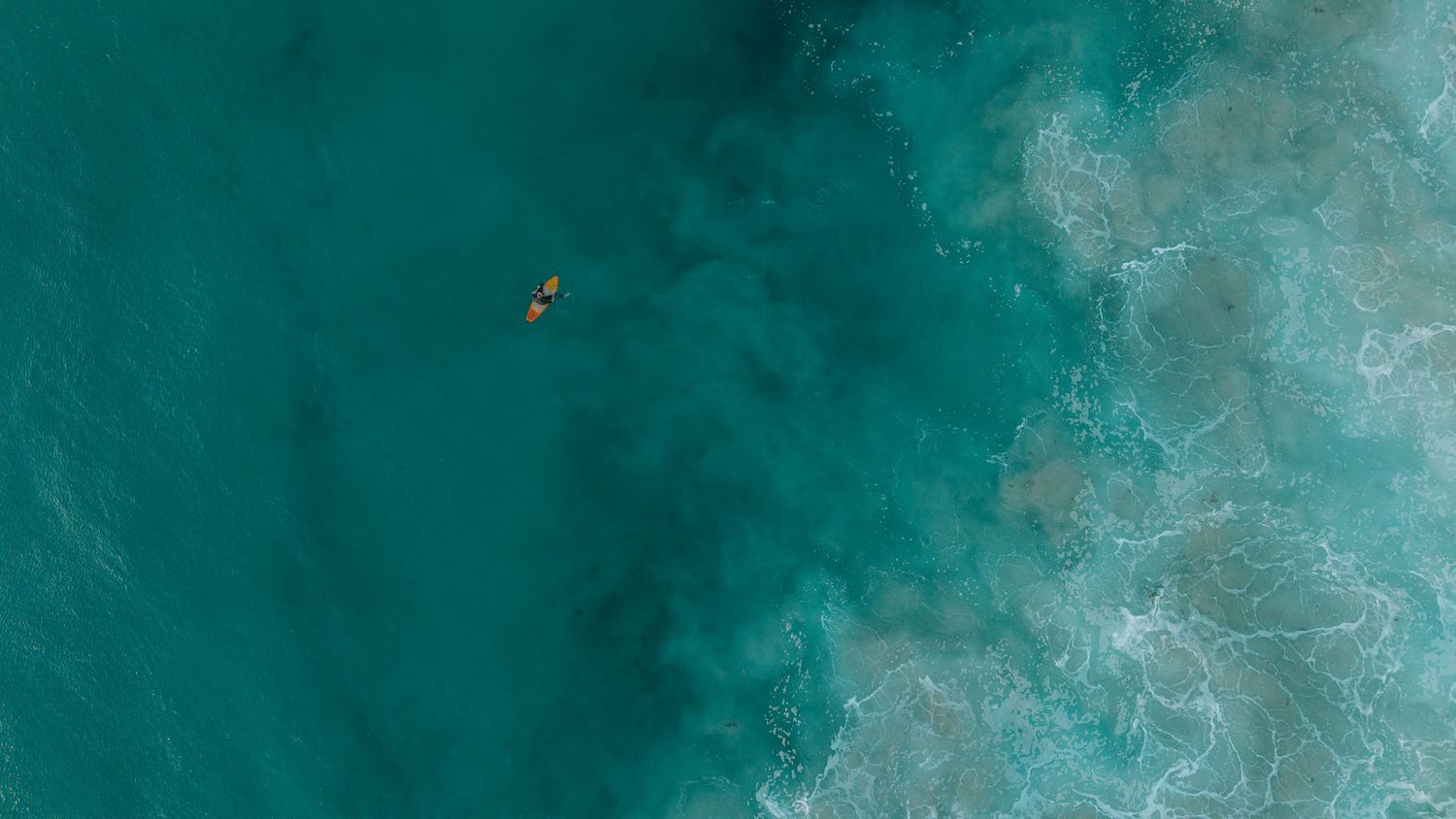 Aerial view of a lone surfer paddling on turquoise ocean waters near a foamy shoreline.