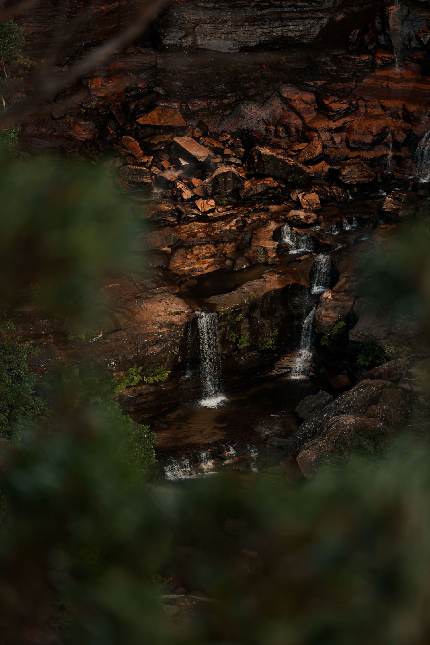 Multiple small waterfalls cascade down dark rocky cliffs in moody forest setting.