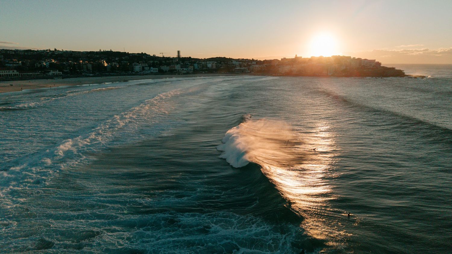 Aerial view of ocean waves rolling toward a sandy beach at golden sunset with a coastal town glowing in the background.