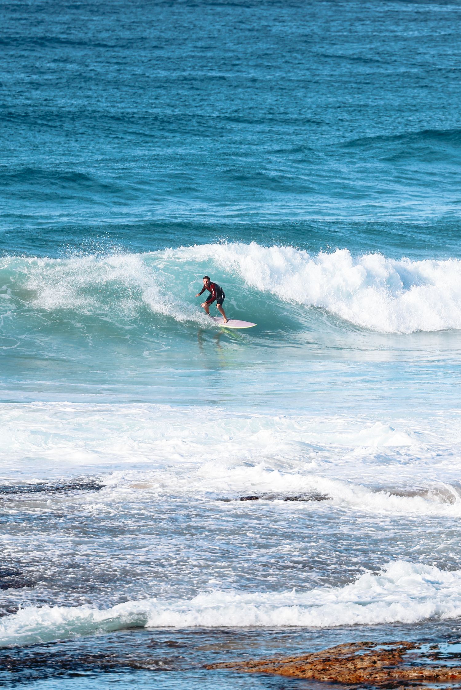 A surfer rides a wave on a white surfboard in clear blue ocean waters near a rocky shoreline.