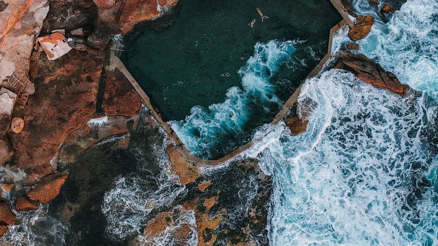 Aerial view of turquoise ocean waves crashing against a rocky coastline with reddish cliffs.
