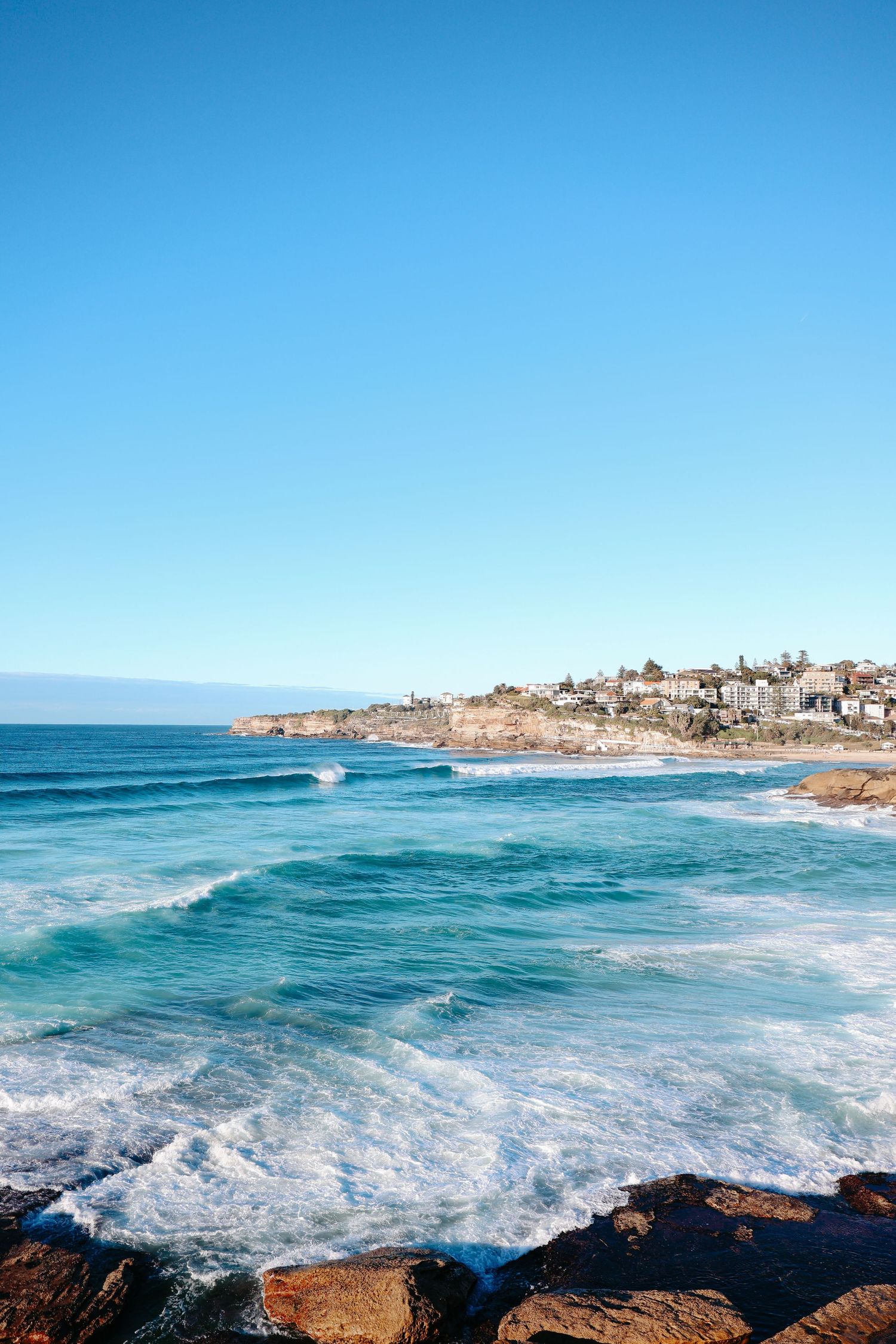 Rocky shoreline with turquoise ocean waves and a coastal town on sandstone cliffs under a clear blue sky.