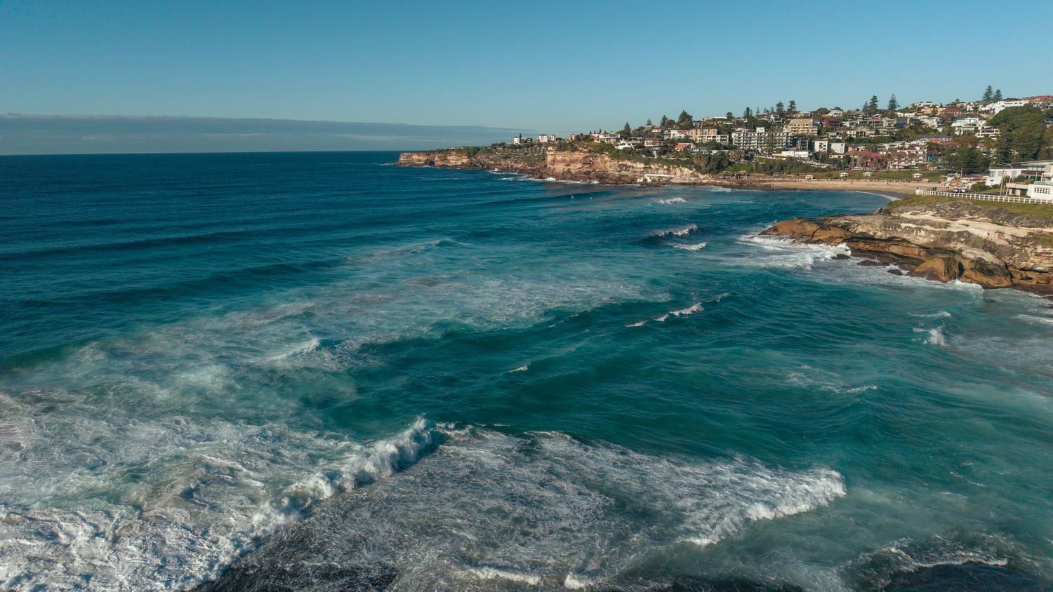 Aerial view of turquoise ocean waves crashing along a rocky Sydney coastline with a residential suburb visible in the background.