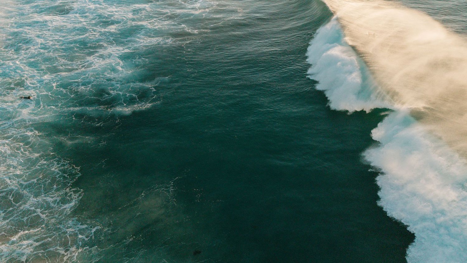 Aerial drone view of powerful ocean waves crashing on sandy beach with deep teal blue water.