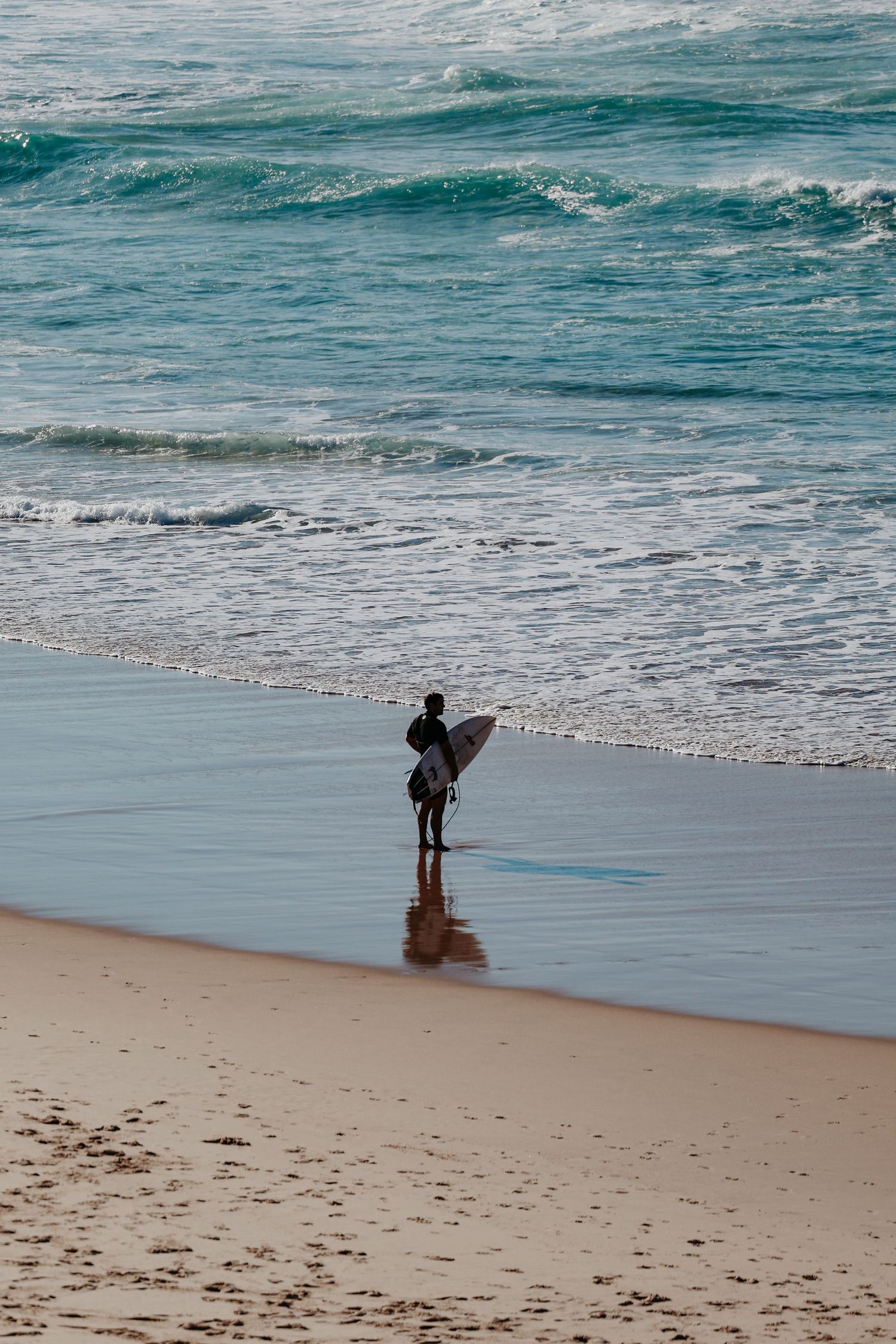 Lone surfer carrying a white surfboard stands at the shoreline of a sandy beach with turquoise ocean waves.