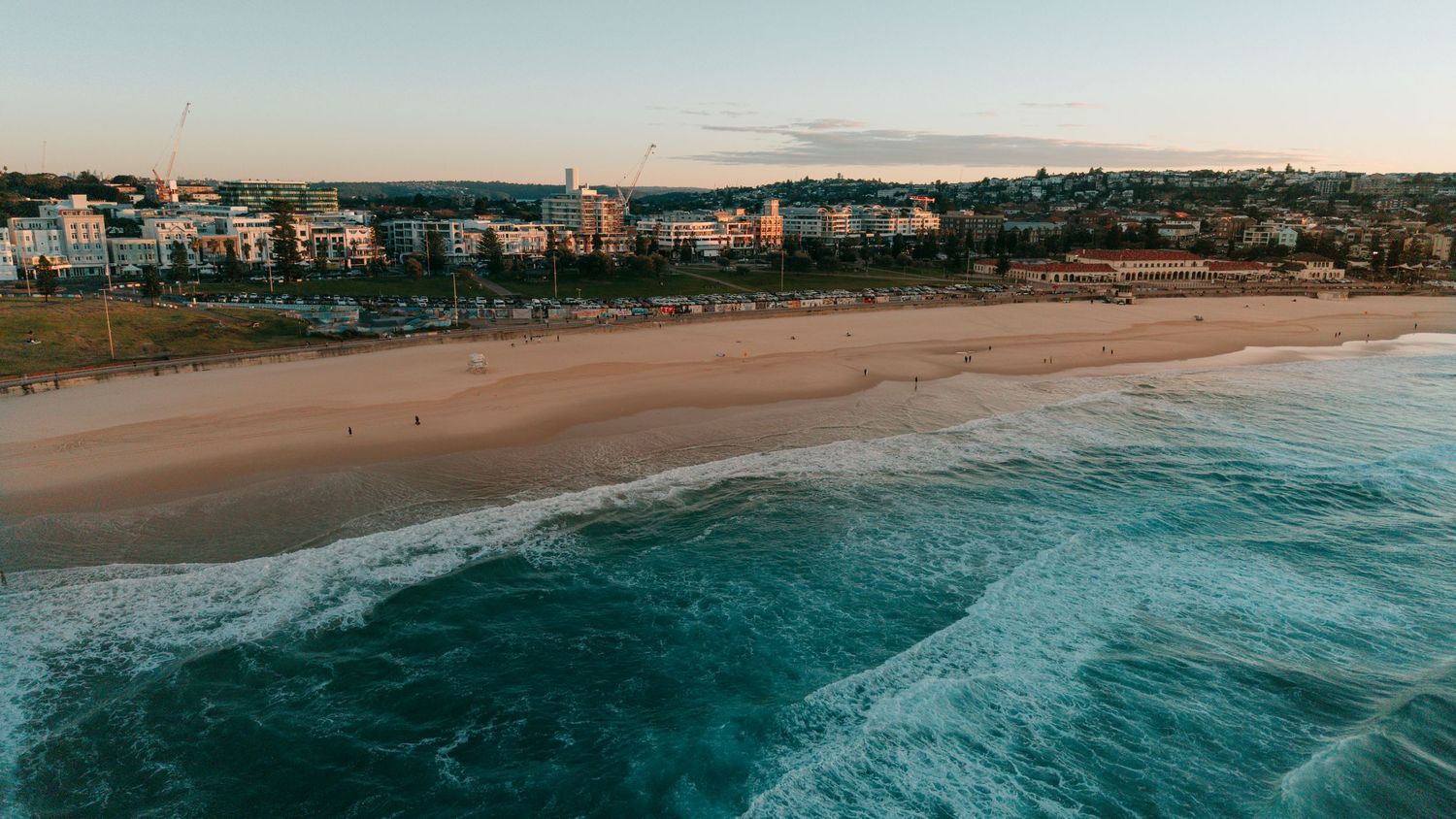 Aerial view of Bondi Beach at sunset with turquoise waves, golden sand, and coastal buildings lining the shoreline.