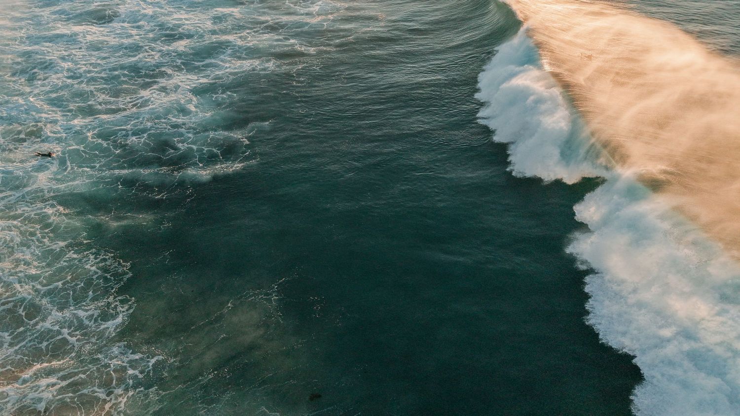Aerial view of ocean waves crashing against a sandy shore, showing contrast between turquoise water and white foam.