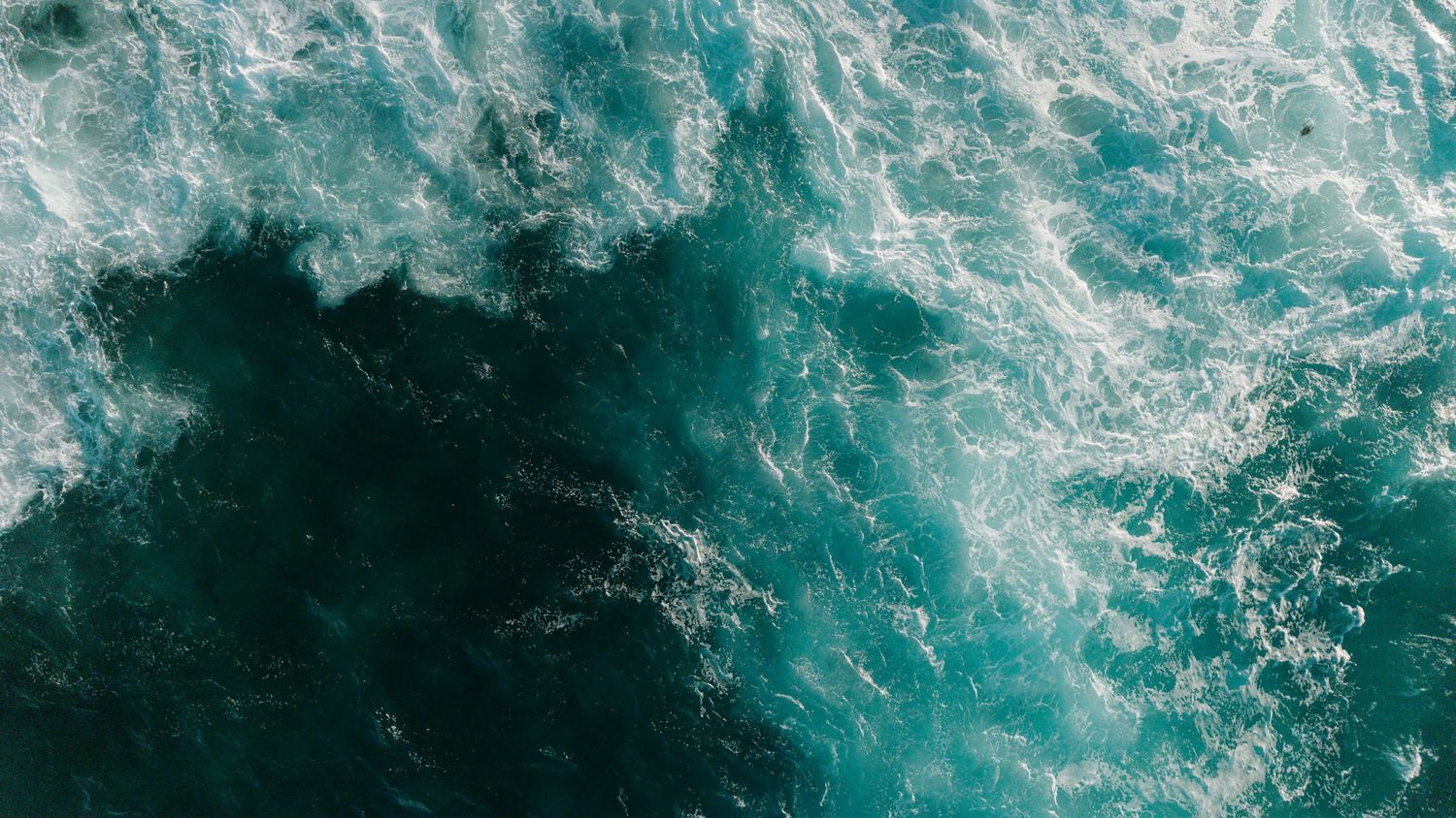 Aerial view of turbulent teal ocean waves with white sea foam swirling on the water surface.