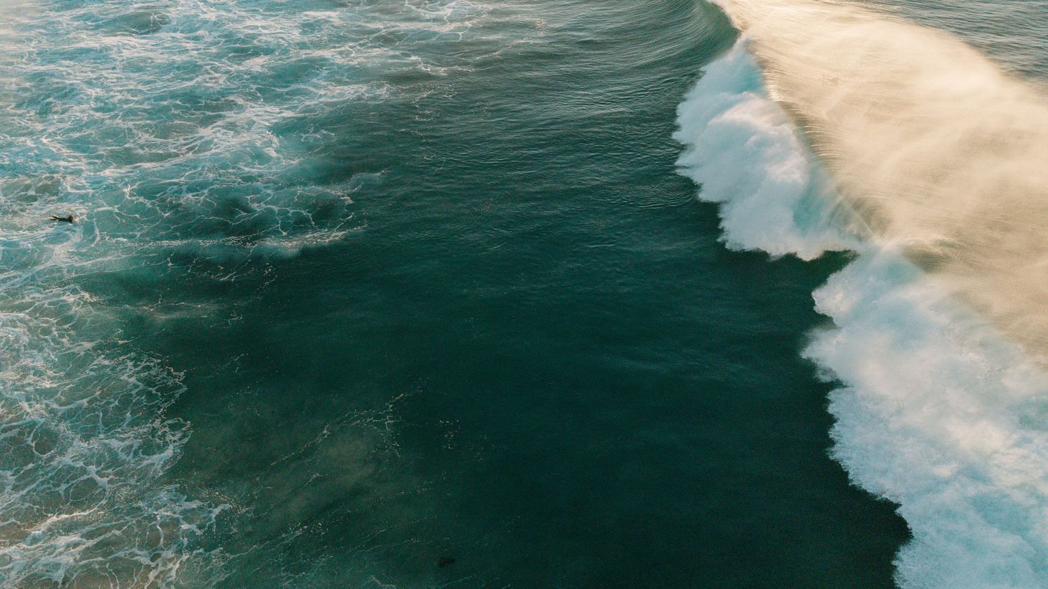 Aerial view of powerful ocean waves crashing onto a sandy shore, showcasing deep teal waters meeting white surf.