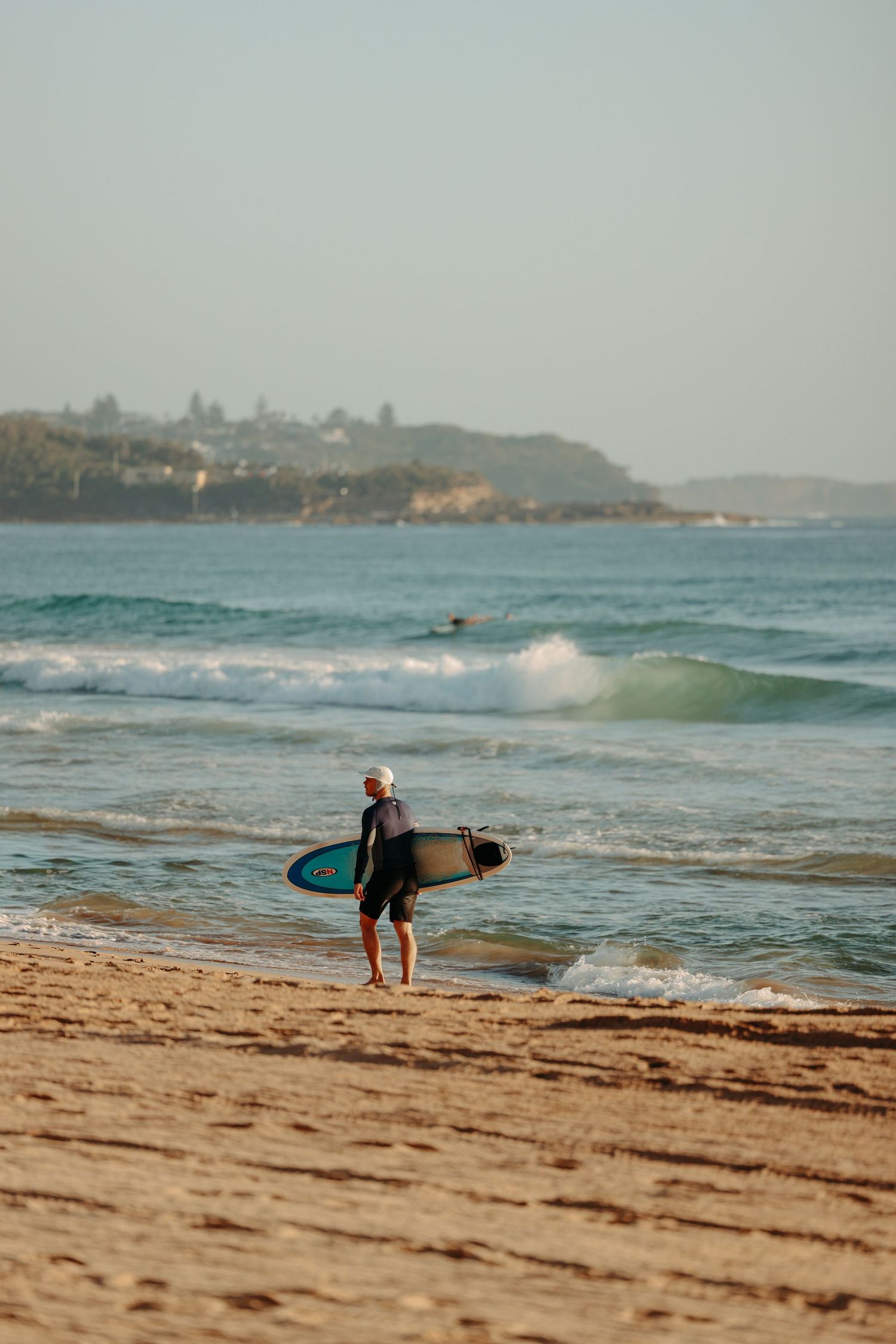 Surfer walking along a sandy beach with surfboard during golden hour.