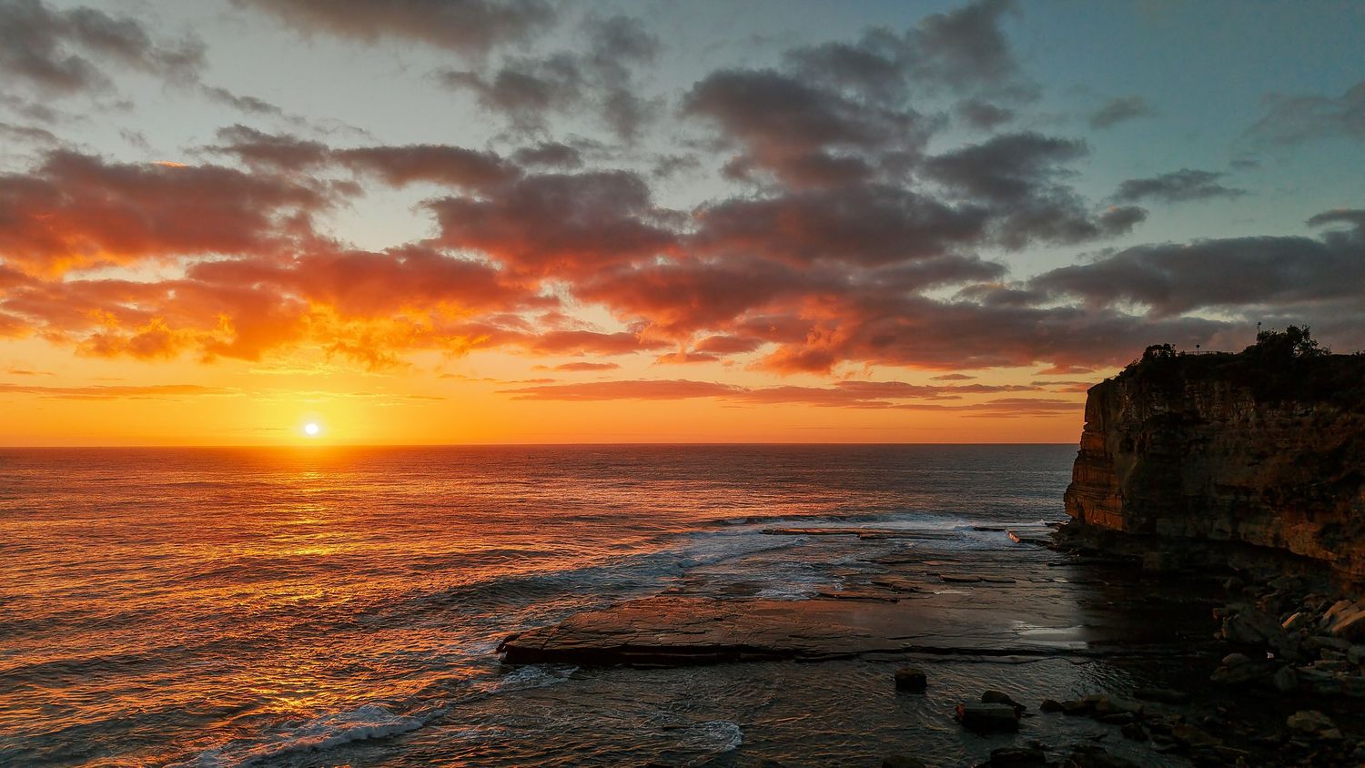 Dramatic sunrise panorama over coastal cliffs with vibrant orange and purple clouds reflecting on the ocean.