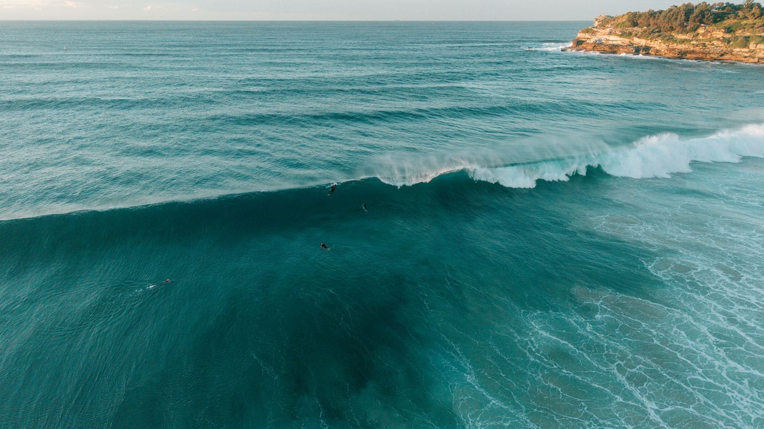 Aerial view of a large turquoise ocean wave breaking near a rocky coastline on a clear day.